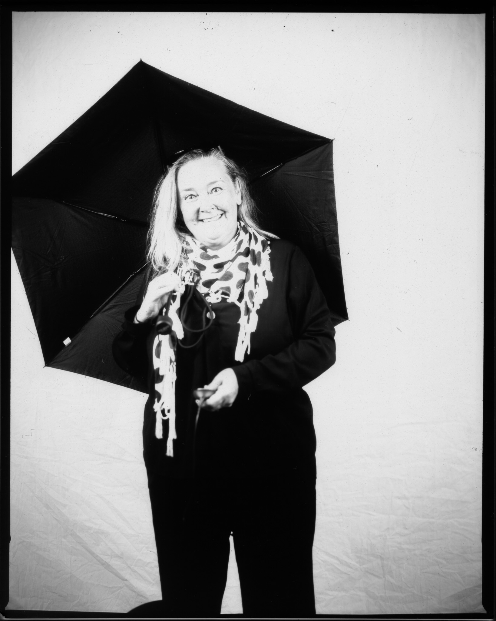 Black and white image of a standing adult smiling holding an open umbrella in front of a white sheet pressing a remote.