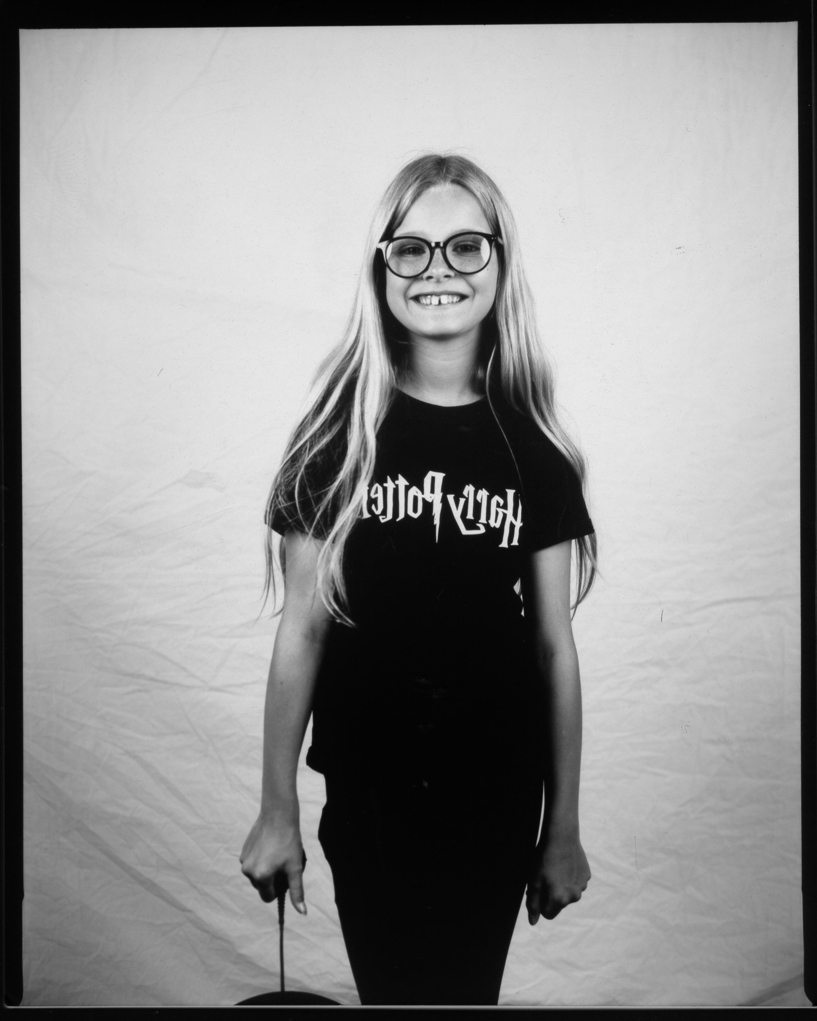 Black and white image of a child smiling in front of a white sheet, pressing a remote with a wire trailing.
