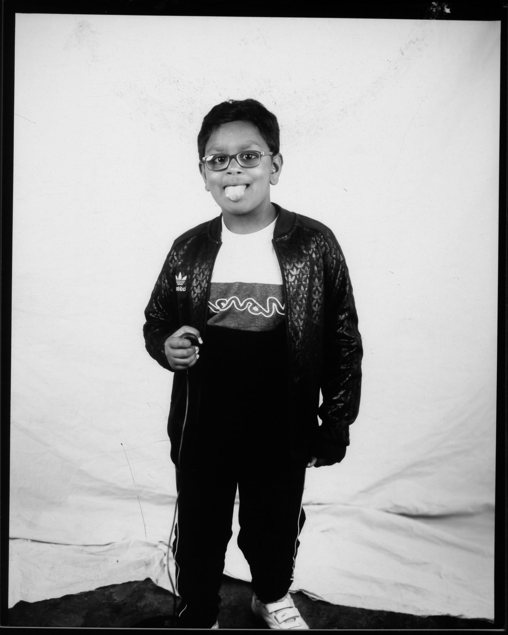 Black and white image of a child with their tongue stuck out in front of a white sheet pressing a remote with wire trailing.