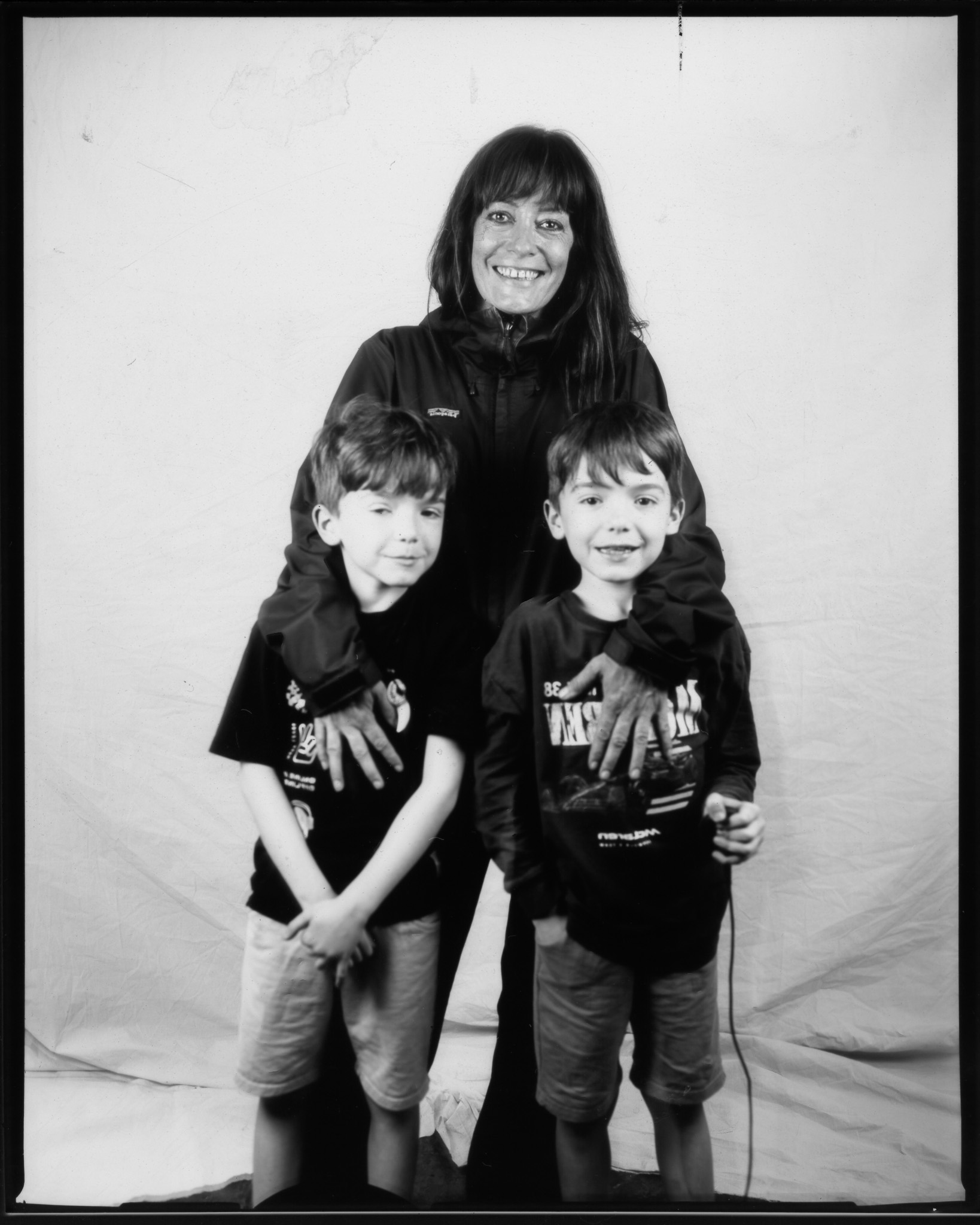 Black and white image of a smiling adult with arms around two children - one presses a remote with long wire.