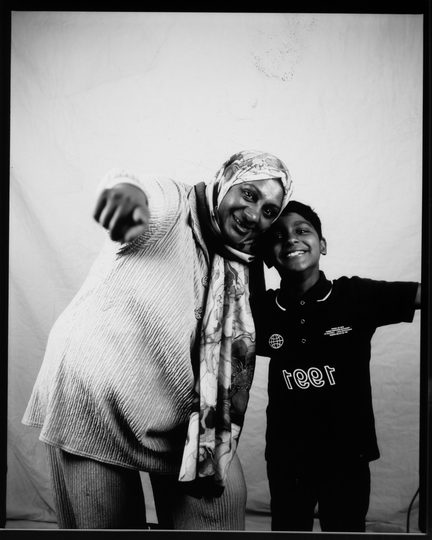 Black and white image of an adult pointing to the camera, a child at their side with arm outstretched, both smiling.