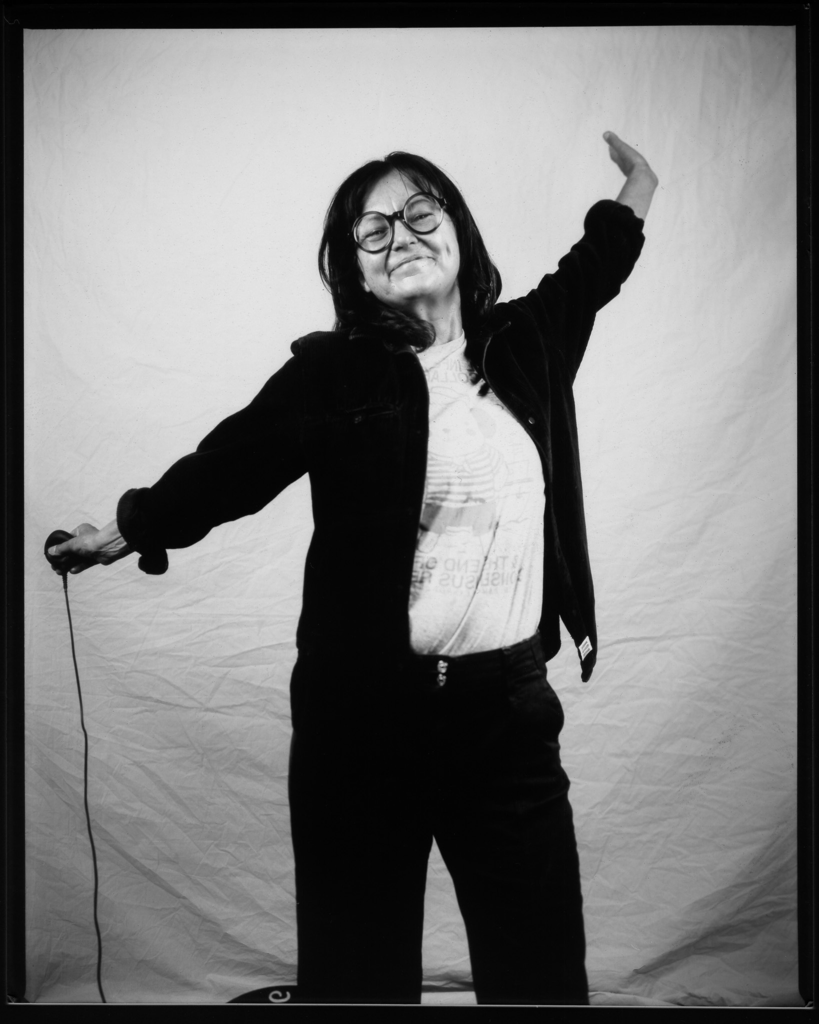 A person with glasses joyfully poses with arms outstretched, against a plain backdrop in a black and white photo.