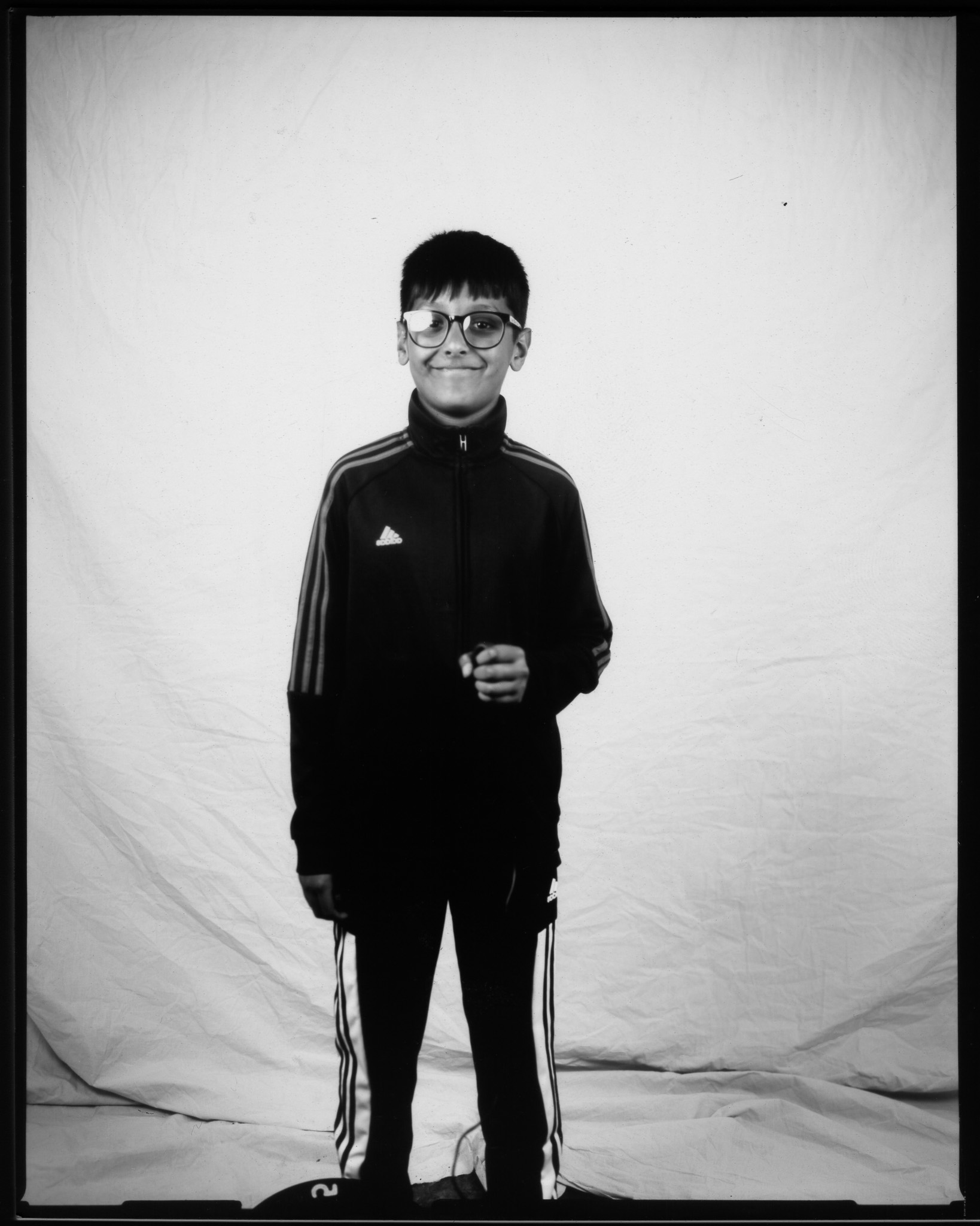 Yes — we can make it smoother and less repetitive. Child wearing glasses and a dark Adidas tracksuit, smiling and posing in a black and white photo.