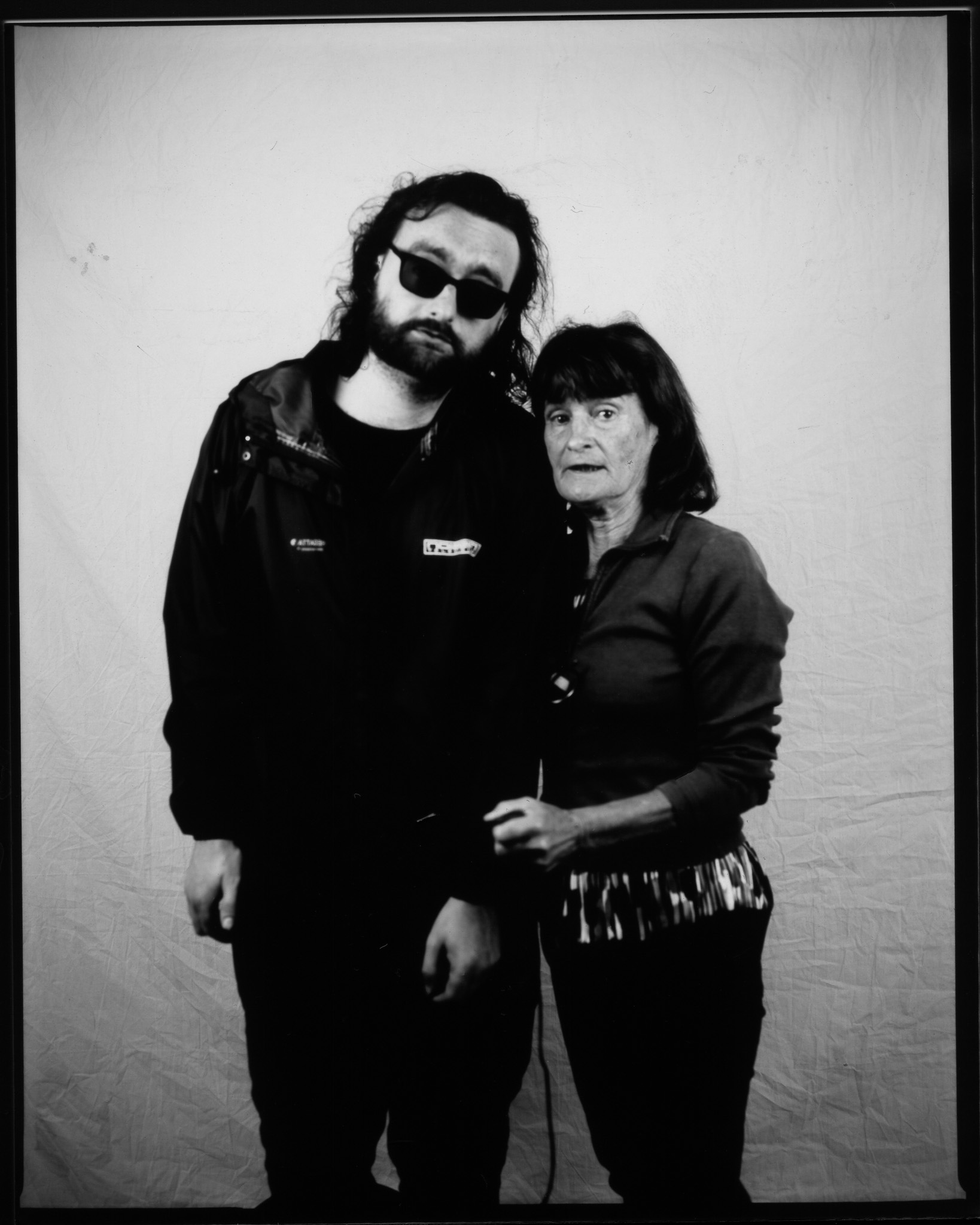 Two adults posing together in a black and white photo, one with tousled hair wearing sunglasses, the other shorter with a fringe, both with neutral expressions.