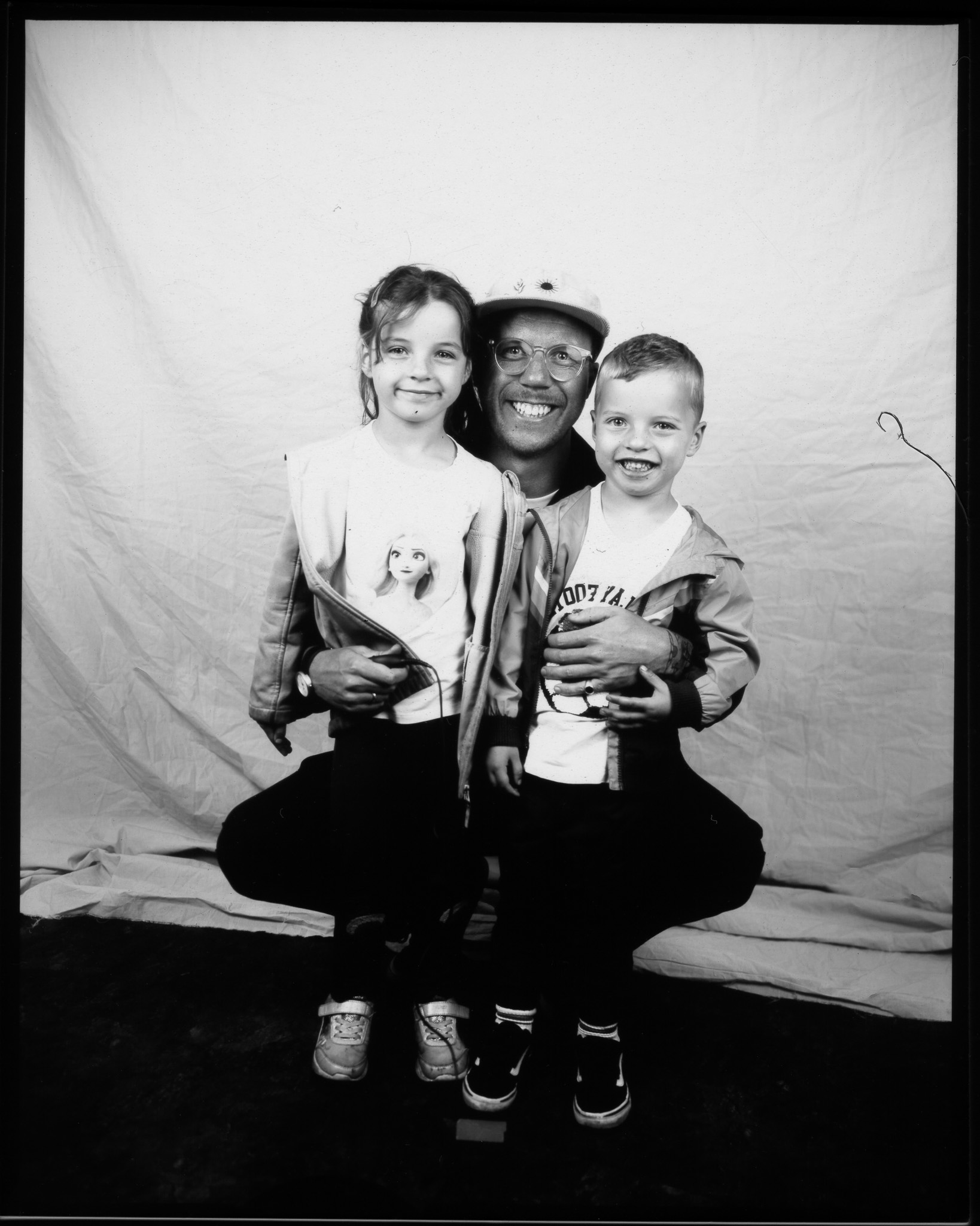 Black and white image of a kneeling adult holding two children, all smiling in front of a white sheet, one pressing a remote.