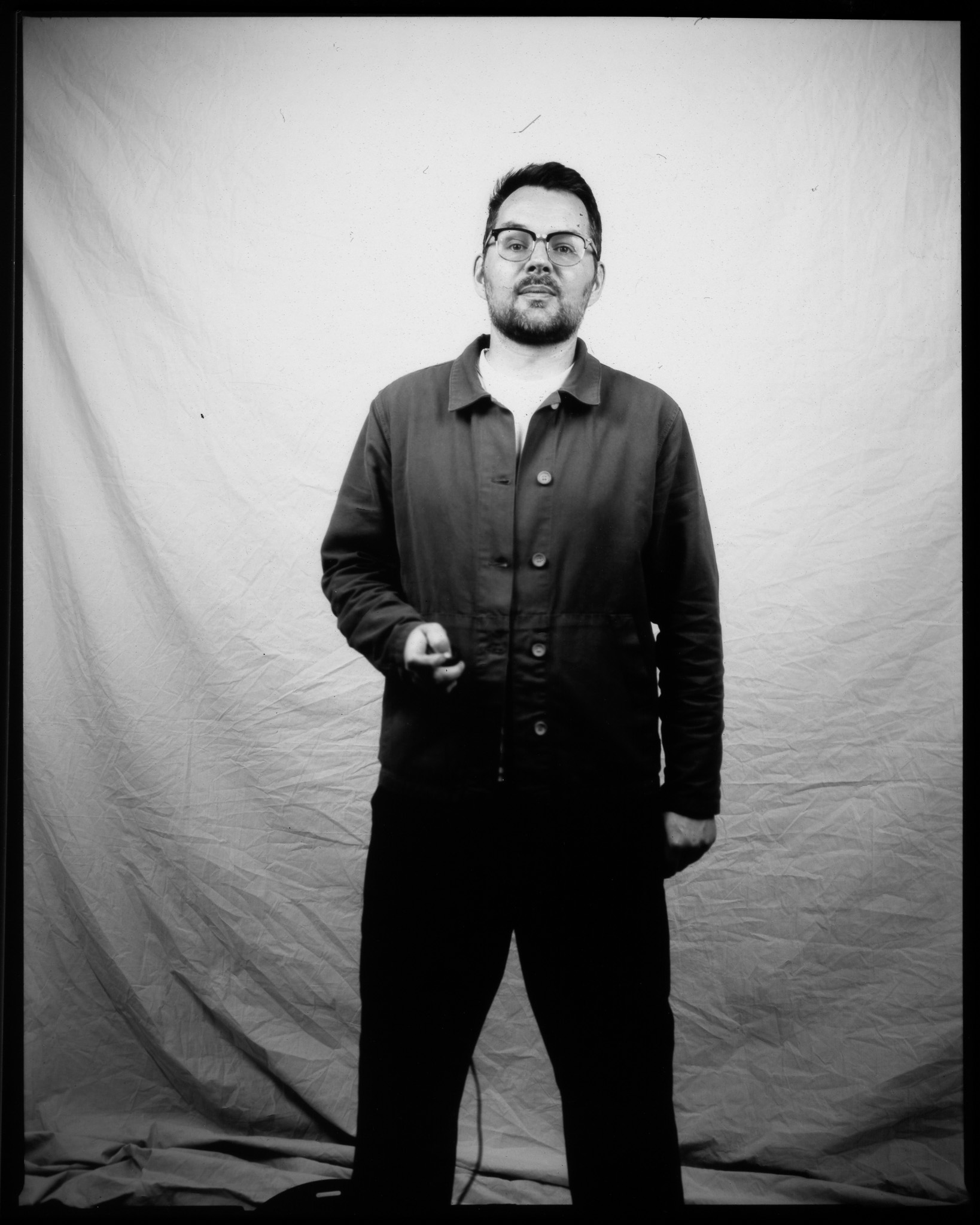 A black and white image of a standing adult in front of a white sheet pressing a remote, a wire trailing to the ground.