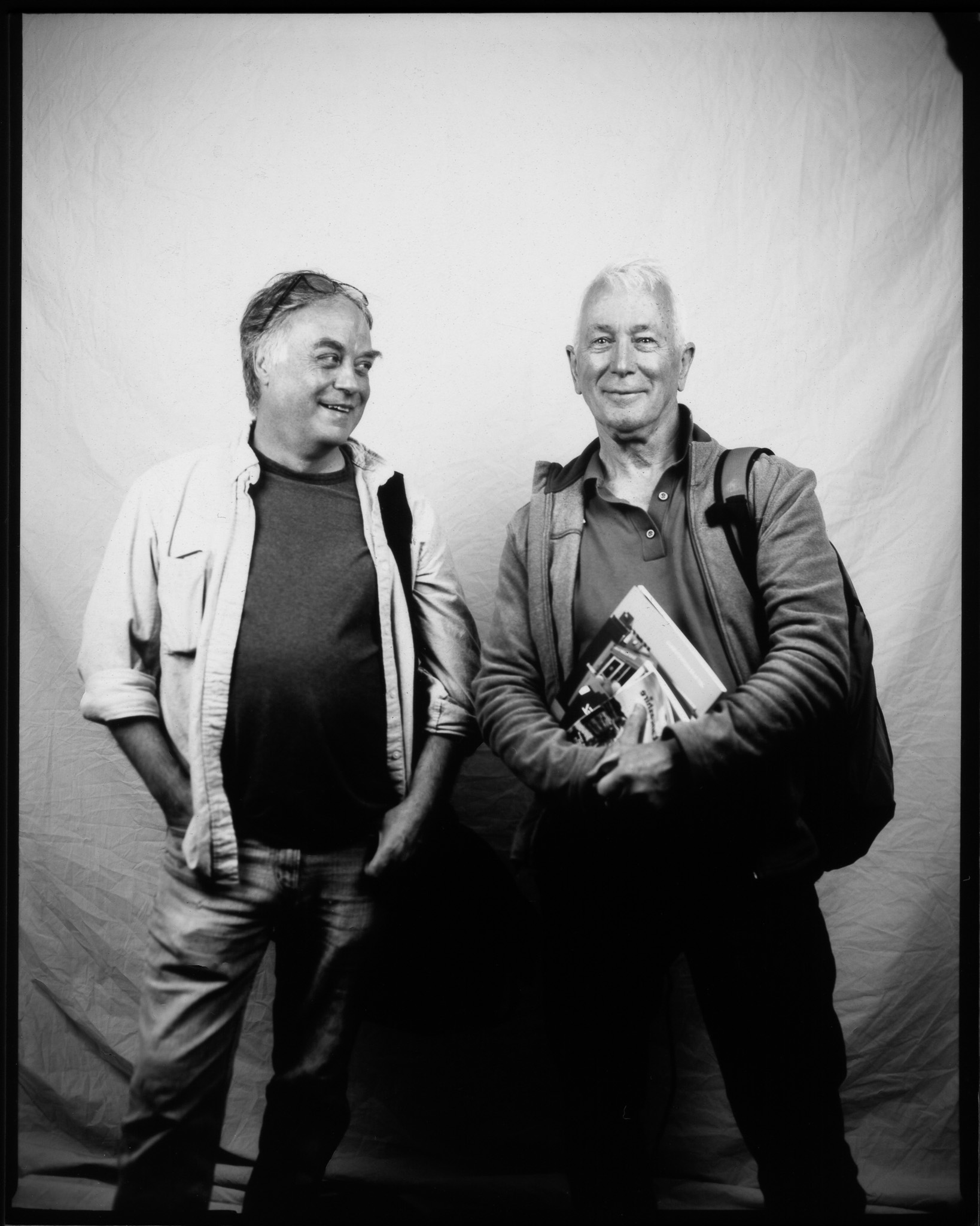 Black and white image of two smiling adults, in front of a white sheet. One looks at the other, who holds a book.