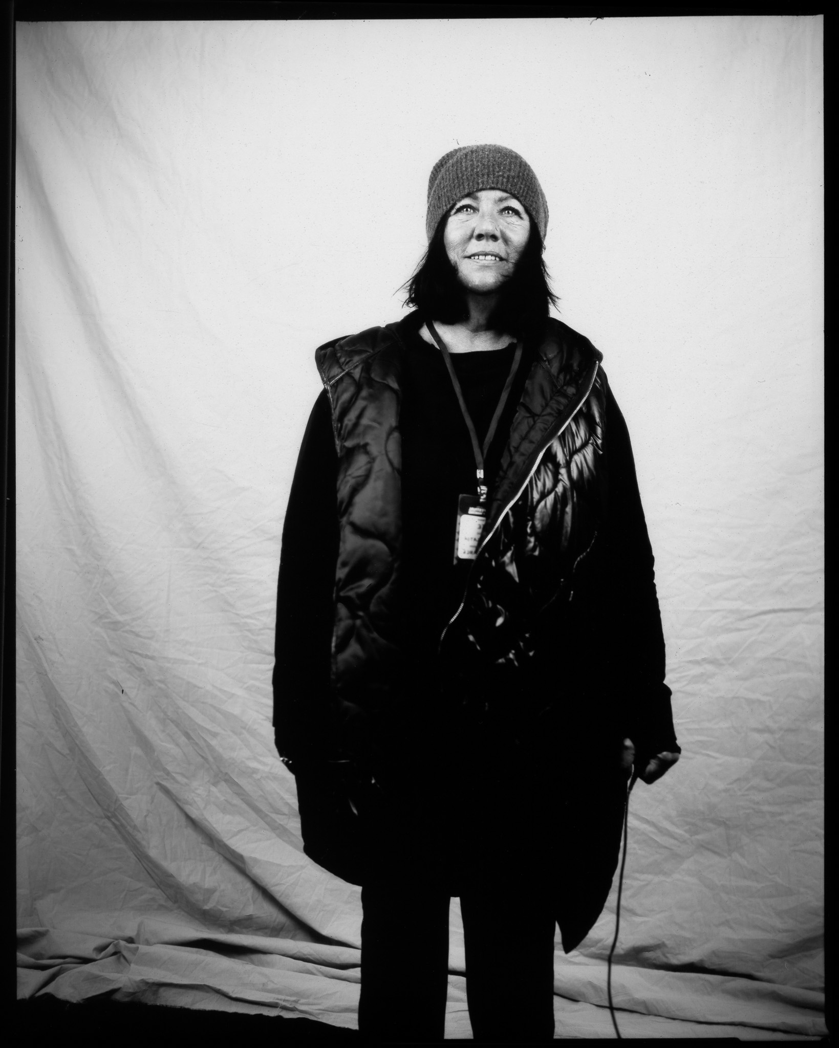 Black and white image of an adult smiling, looking away, in front of a white sheet while pressing a remote, wire trailing.