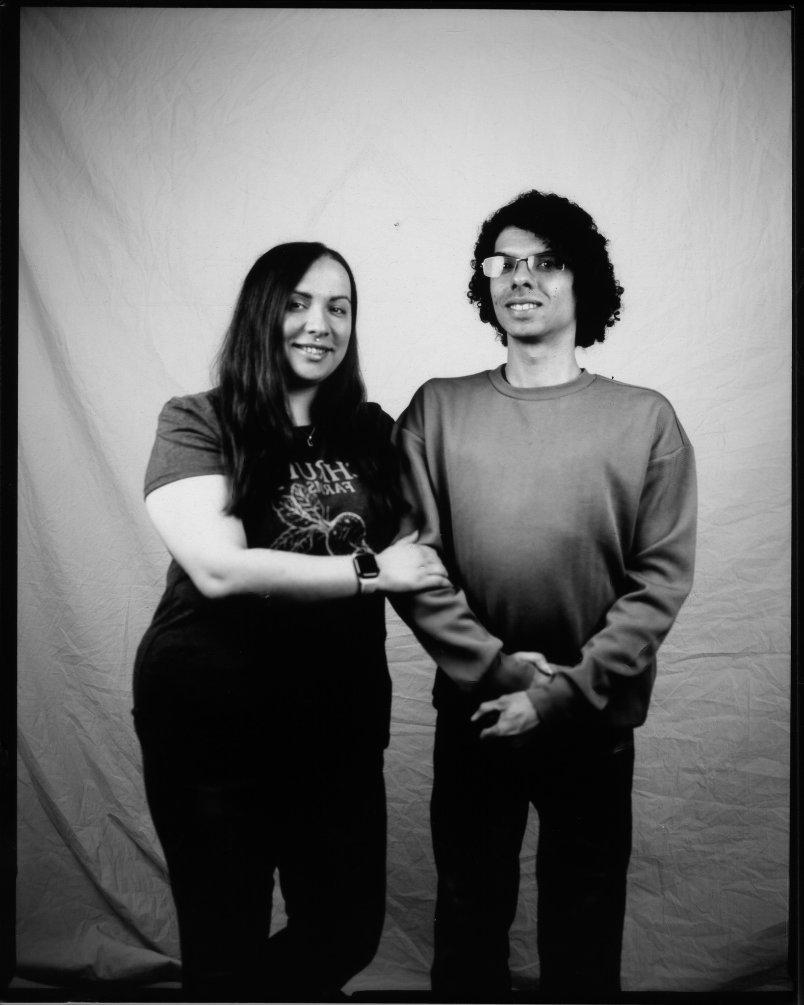 A black and white image of two standing adults, with arms linked smiling in front of a white sheet.