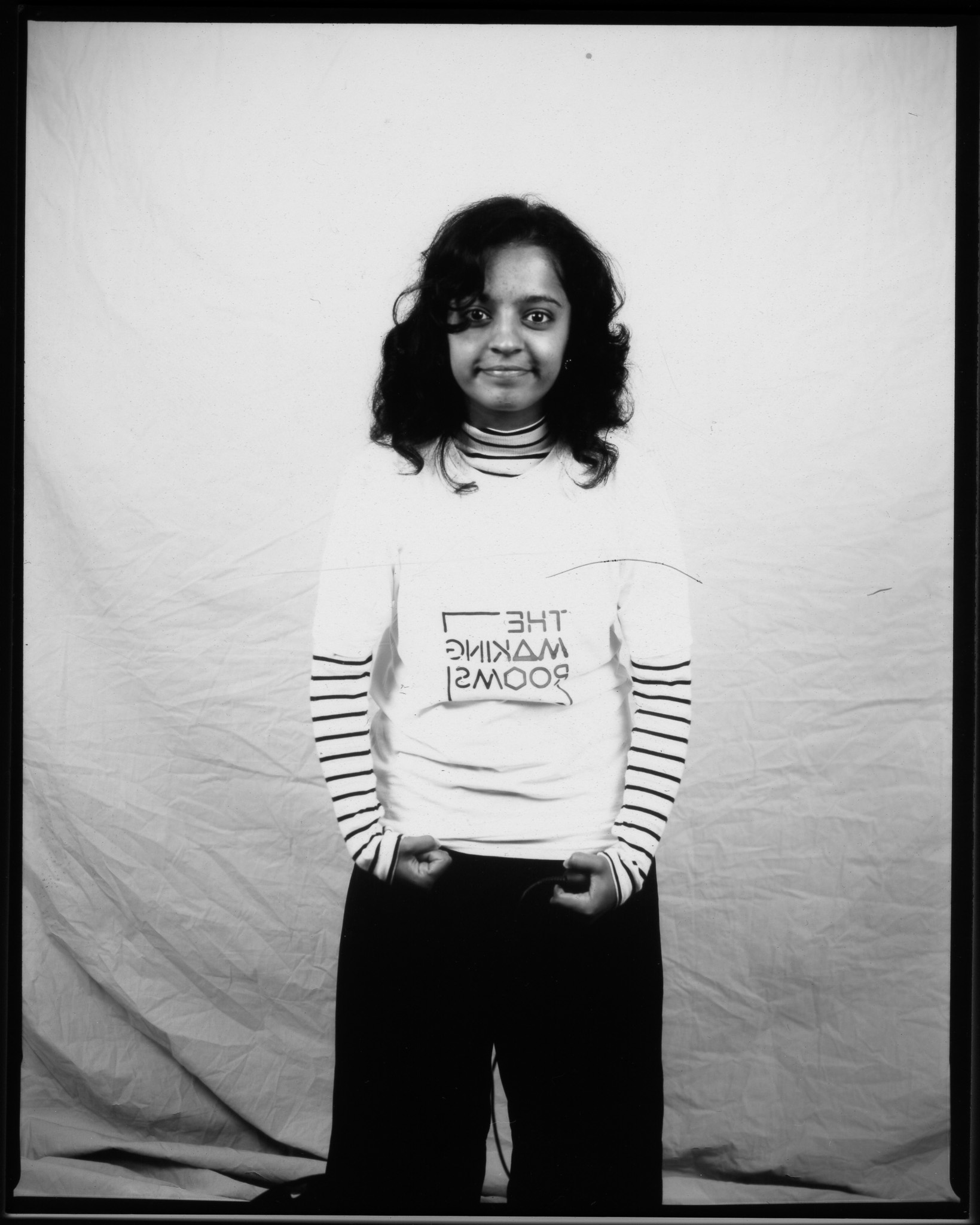 Black and white image of a young person stood smiling in front of a white sheet, holding their trouser loops.
