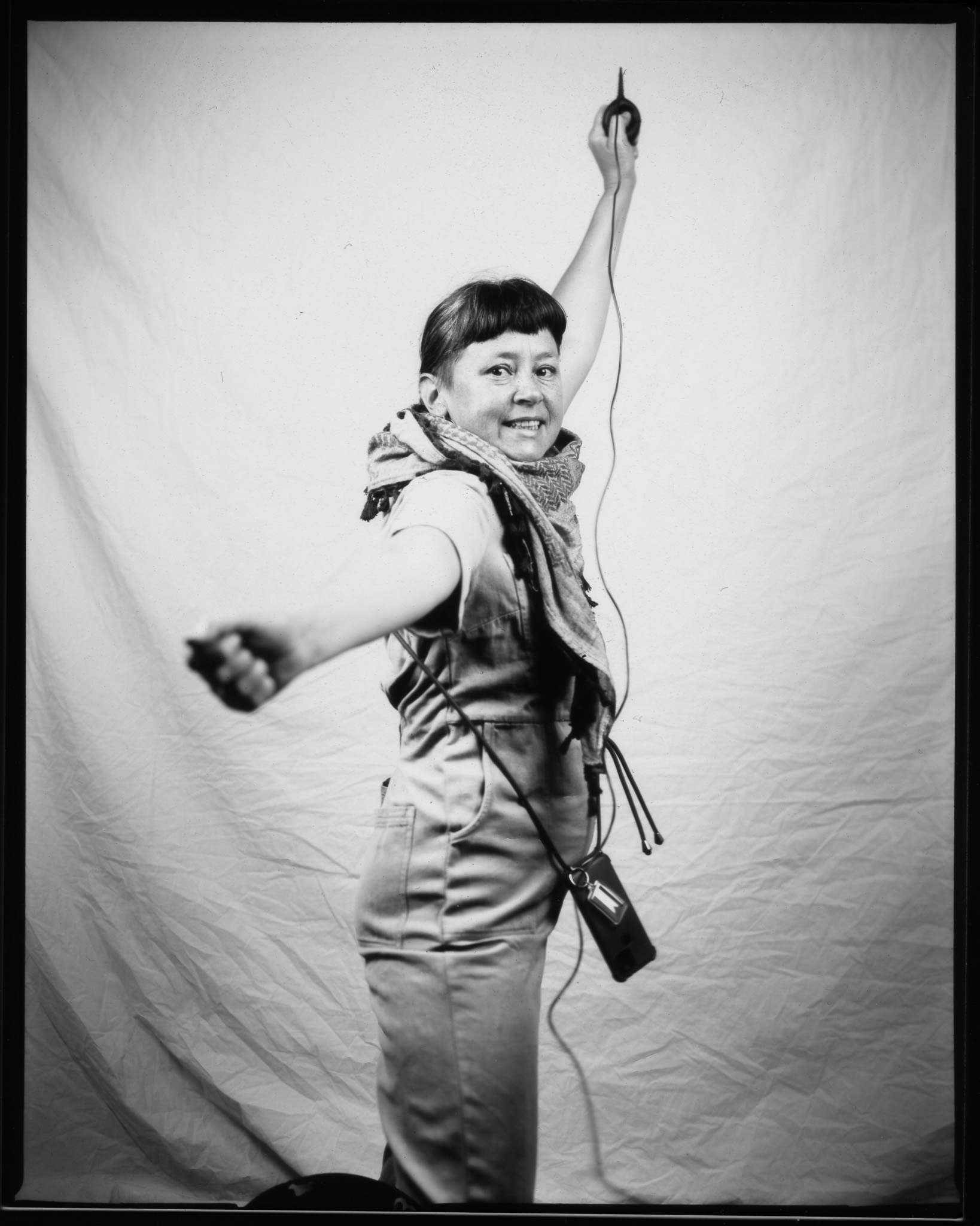 Adult with hair tied up and fringe, standing sideways with arms stretched out and posing in a black and white photo.