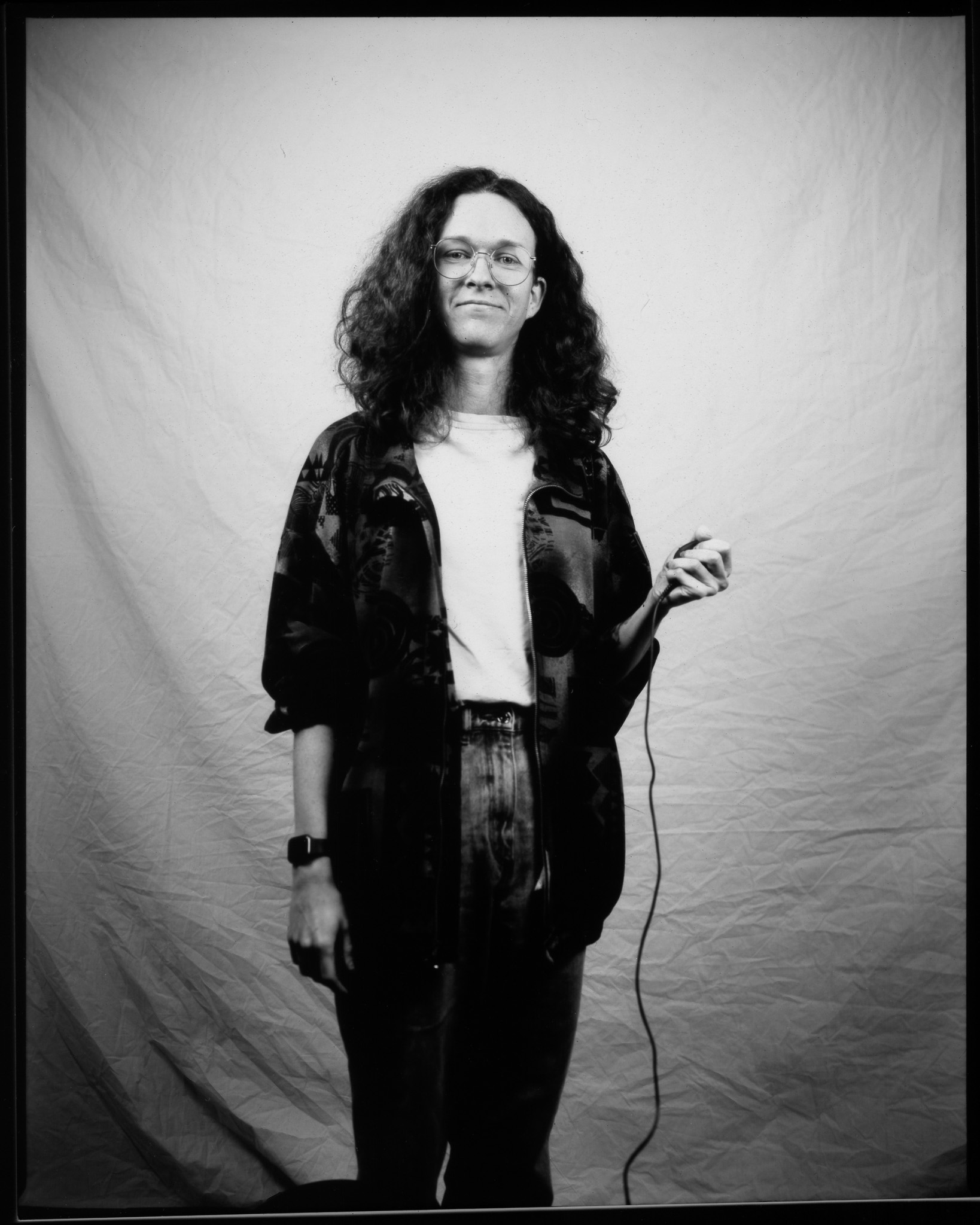 Adult with long curly hair and glasses, wearing a white T-shirt, smiling and posing in a black and white photo.