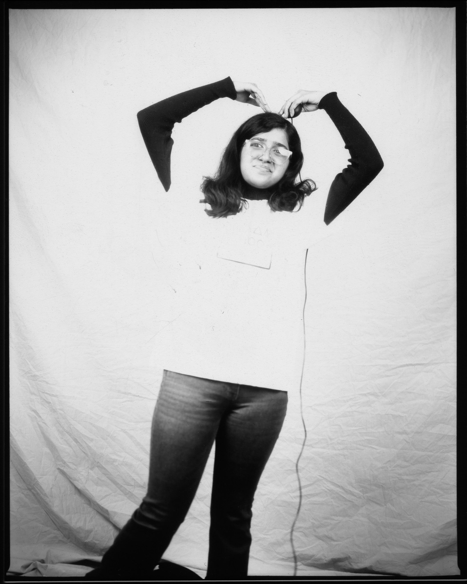 Adult with long hair and glasses, smiling and making a heart shape with their arms, posing in a black and white photo.