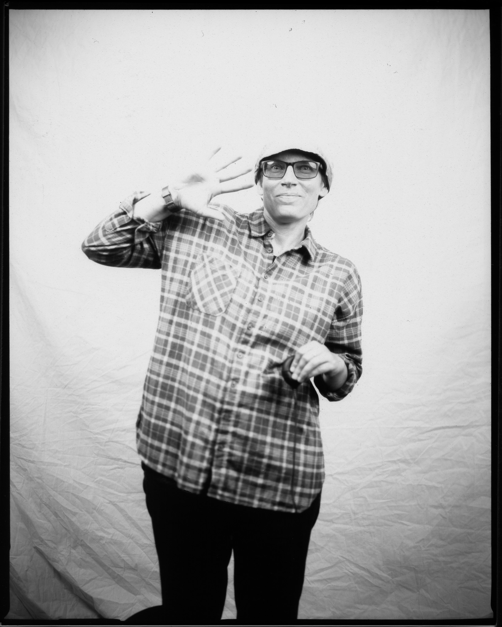 Adult wearing a hat and sunglasses, dressed in a plaid shirt, smiling and waving at the camera and posing in a black and white photo.