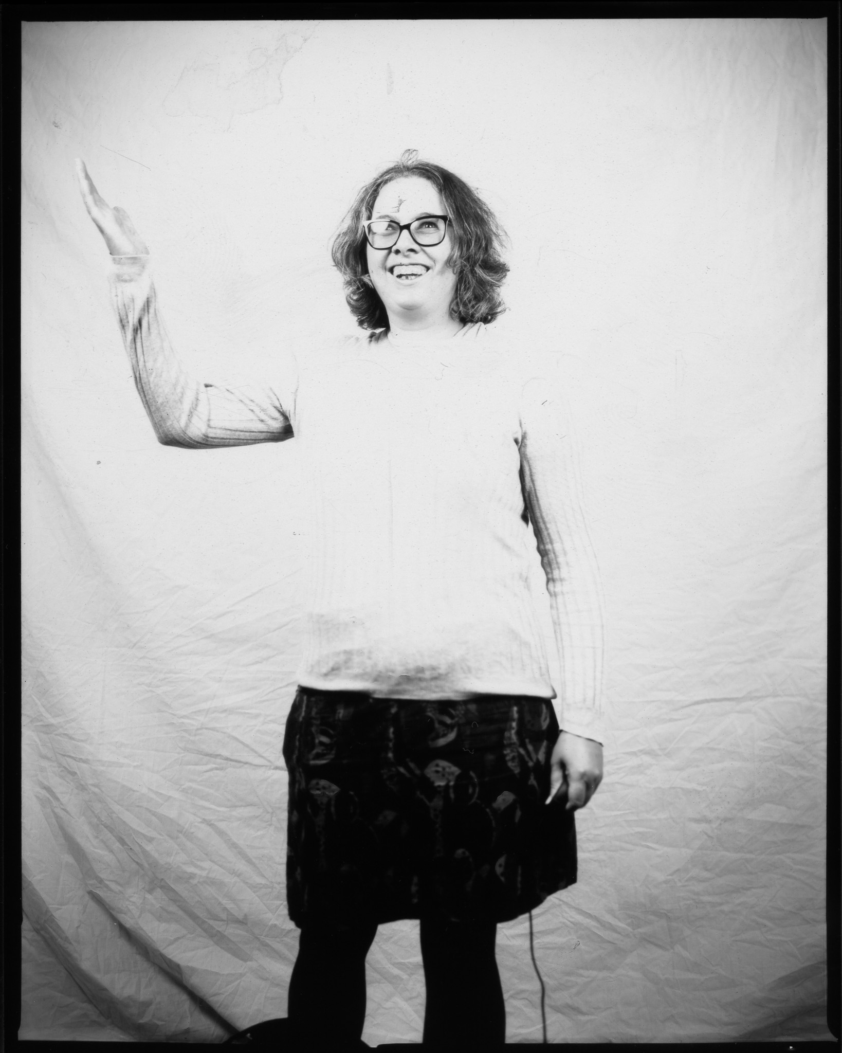 Adult with mid-length hair and glasses, smiling broadly with one hand raised, wearing a jumper and skirt and posing in a black and white photo.