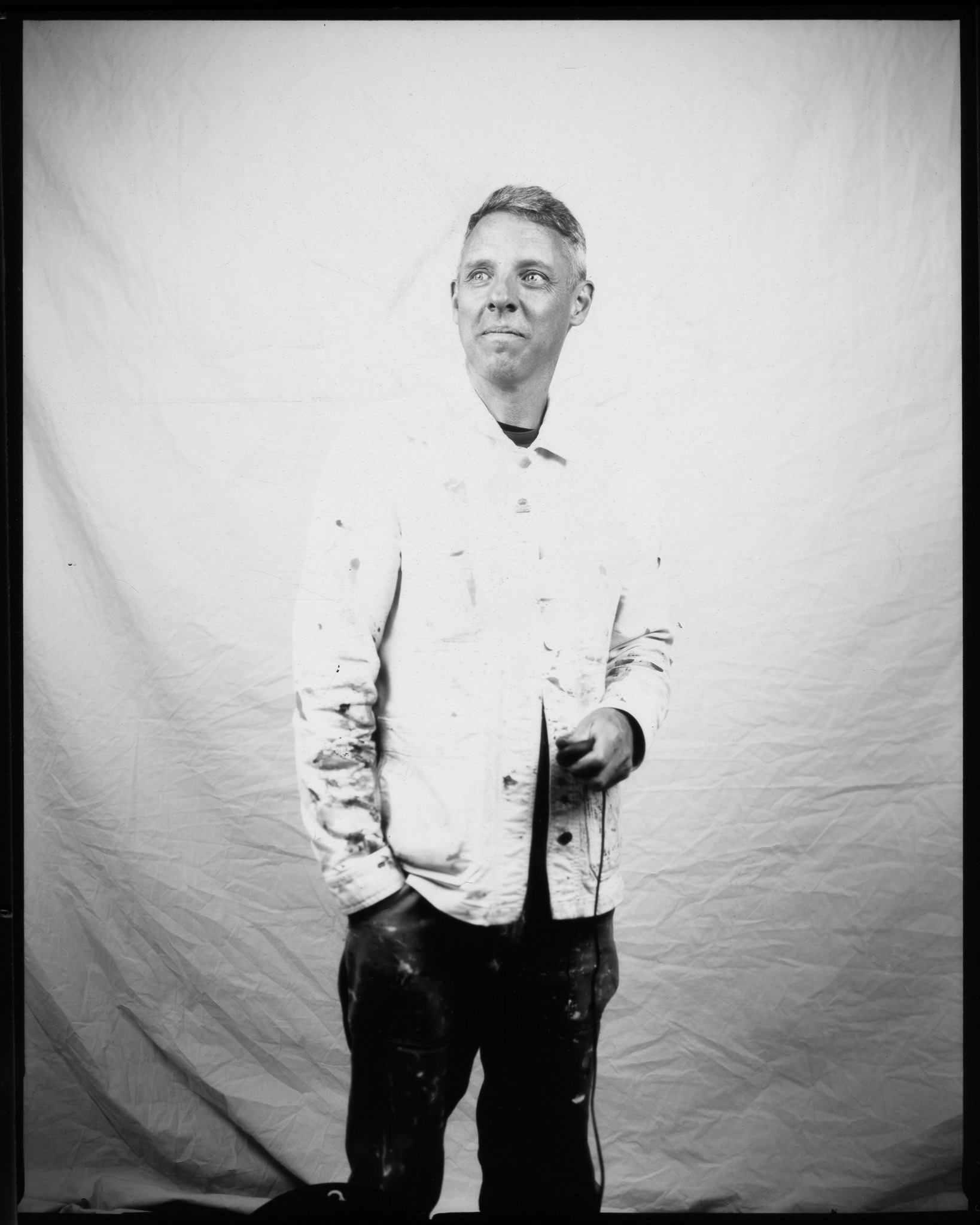 Adult smiling and looking to the side, wearing a paint-covered jacket and posing in a black and white photo.