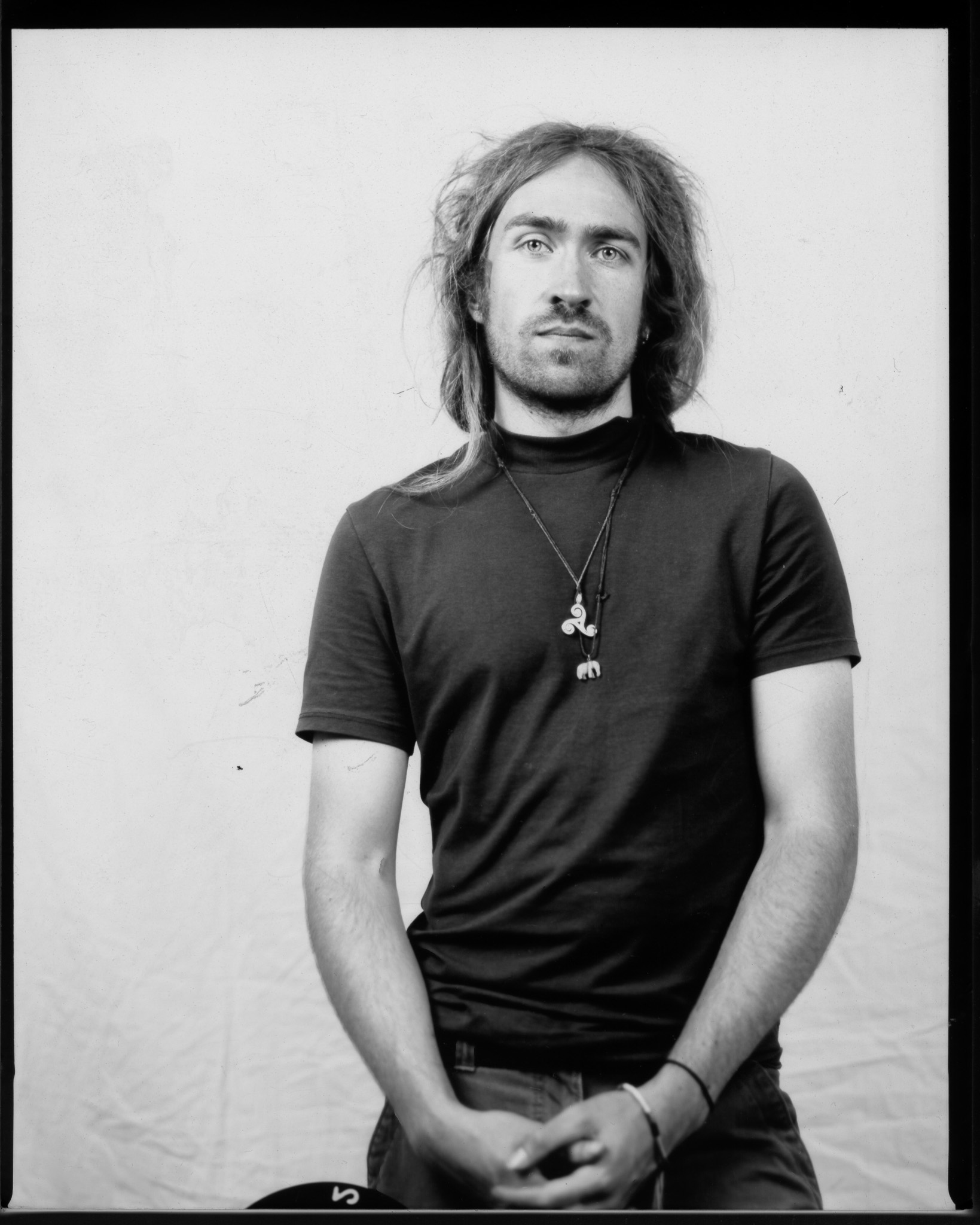 Adult with tousled hair tied back, holding hands in front, wearing long rope necklaces with pendants, and posing with a neutral expression in a black and white photo.