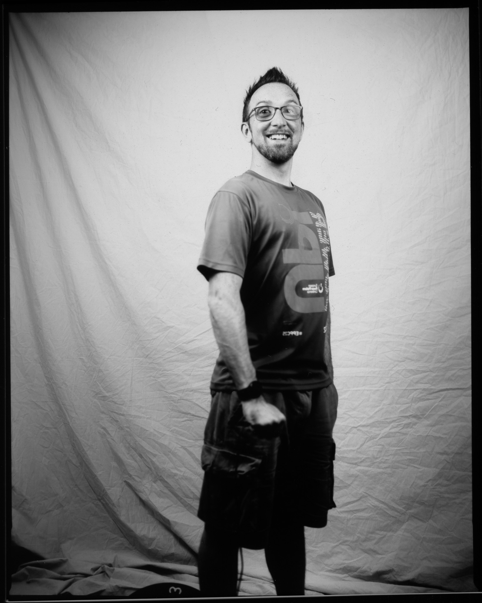 Adult with spiky hair and glasses, wearing a T-shirt and cargo shorts, smiling and posing in a black and white photo.