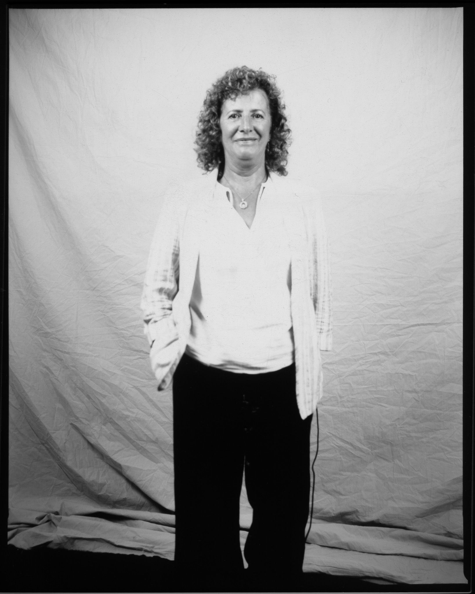 Adult with curly mid-length hair smiling, wearing a striped overshirt and necklace, hand in trouser pocket, posing in a black and white photo.