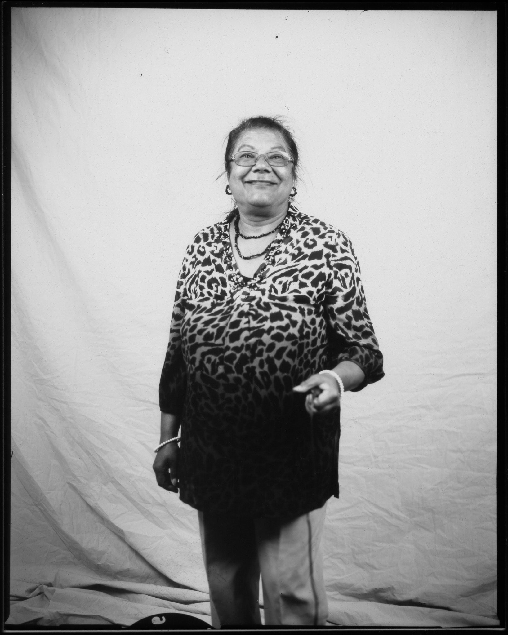 Adult smiling at the camera, hair tied up, wearing glasses, hoop earrings, a beaded necklace, and a leopard print top, posing in a black and white photo.