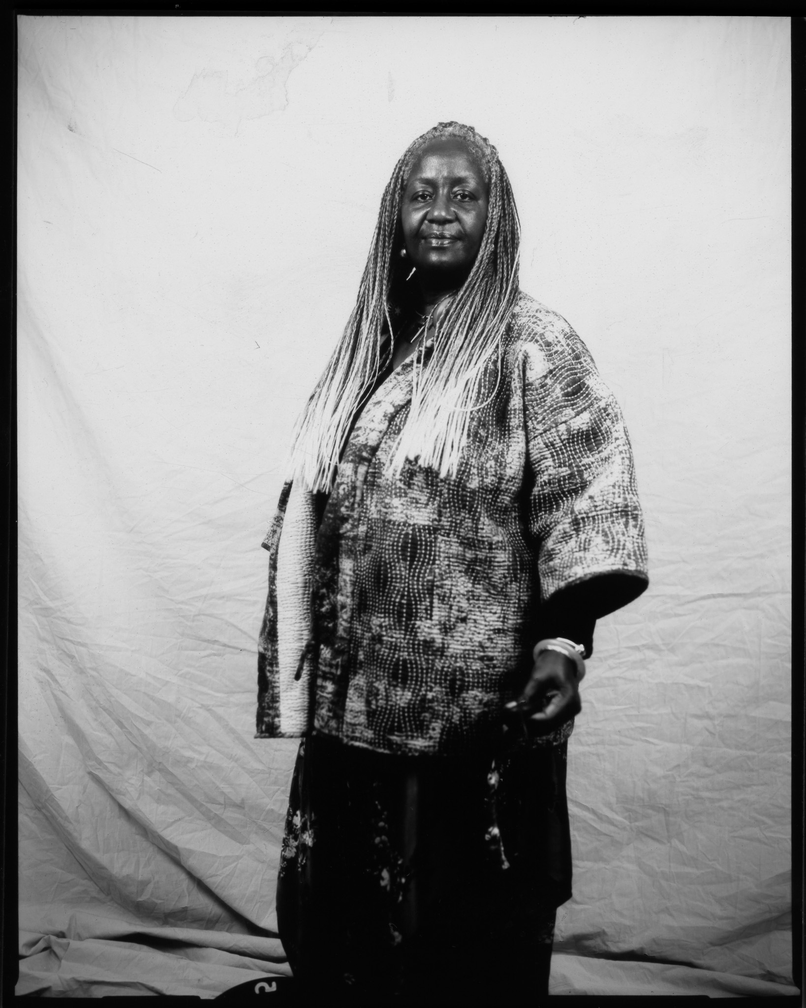 Adult with long braids turned to the side, looking at the camera, wearing a jacket with an abstract pattern and posing in a black and white photo.