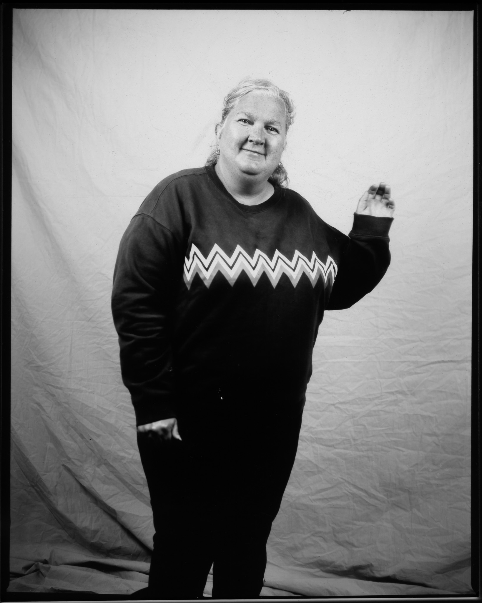 Adult with hair tied back, smiling and waving at the camera, wearing a zig-zag patterned jumper and posing in a black and white photo.