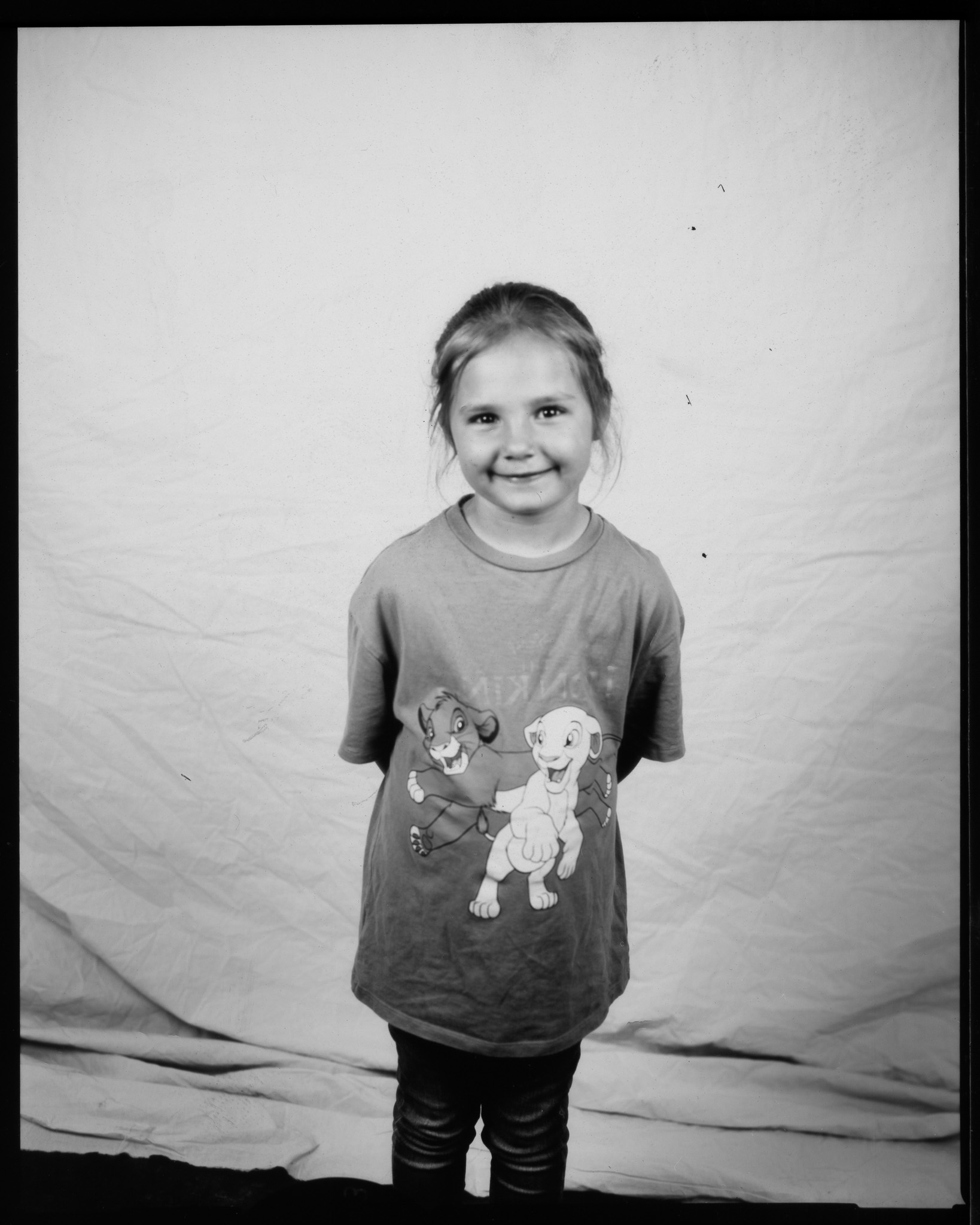 Small child smiling with hands behind their back, wearing a Lion King T-shirt and posing in a black and white photo.