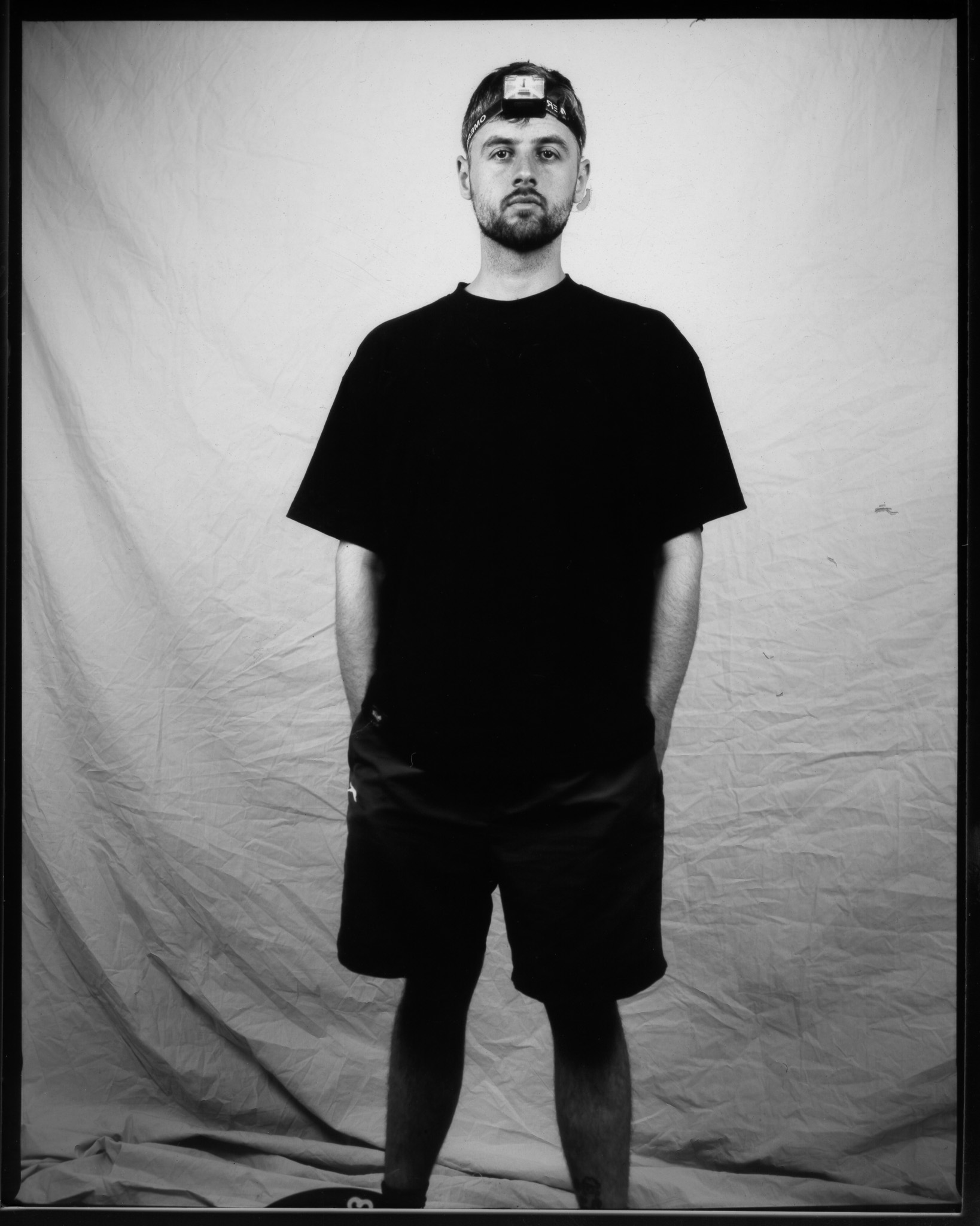 Adult with a neutral expression wearing a black T-shirt, hand in shorts pocket, and a headlamp for taking photos, posing in a black and white photo.
