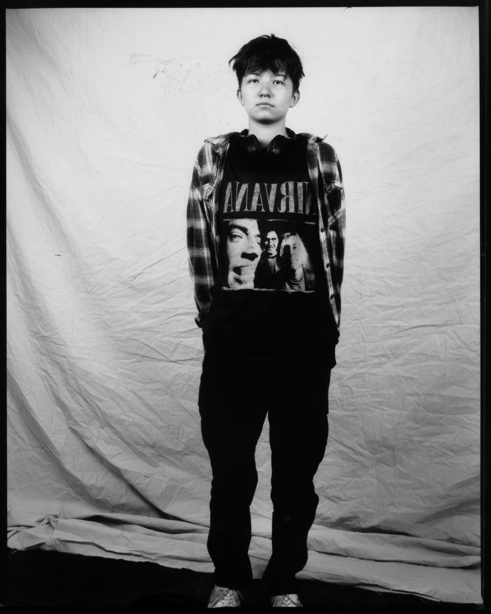 Teenager with short dark hair wearing a plaid shirt over a Nirvana T-shirt, posing with a neutral expression in a black and white photo.