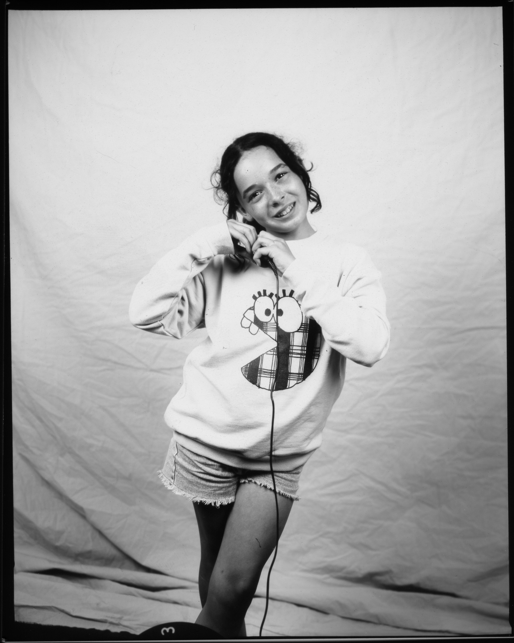 Child smiling with head tilted to the side, wearing a cartoon jumper and denim shorts, making a heart shape with their hands and posing in a black and white photo.