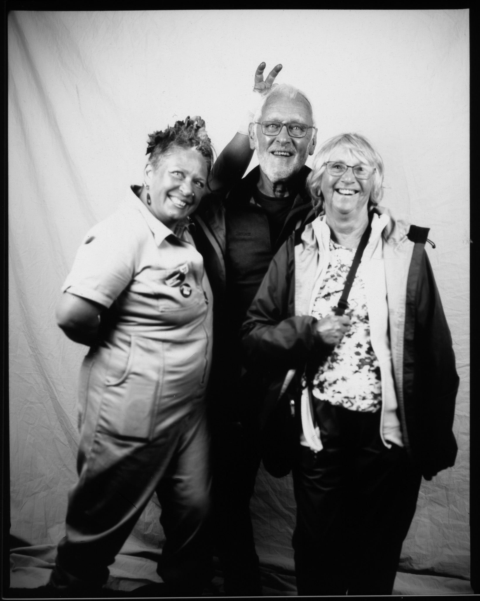 Three people pose playfully against a plain backdrop in a black-and-white photo, smiling, with one person making bunny ears behind another.