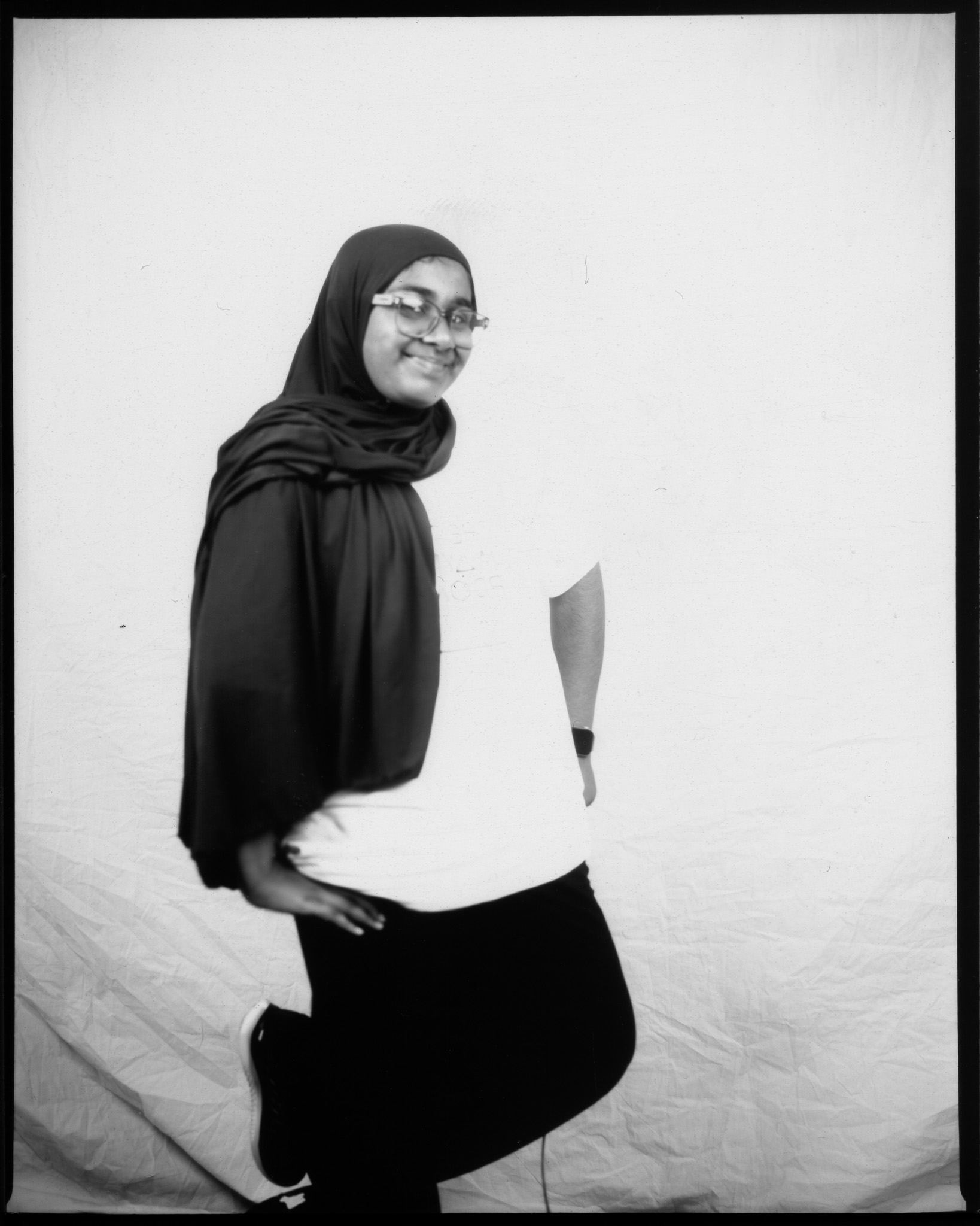 A smiling person in glasses and a hijab poses playfully against a plain backdrop in a black and white photo.