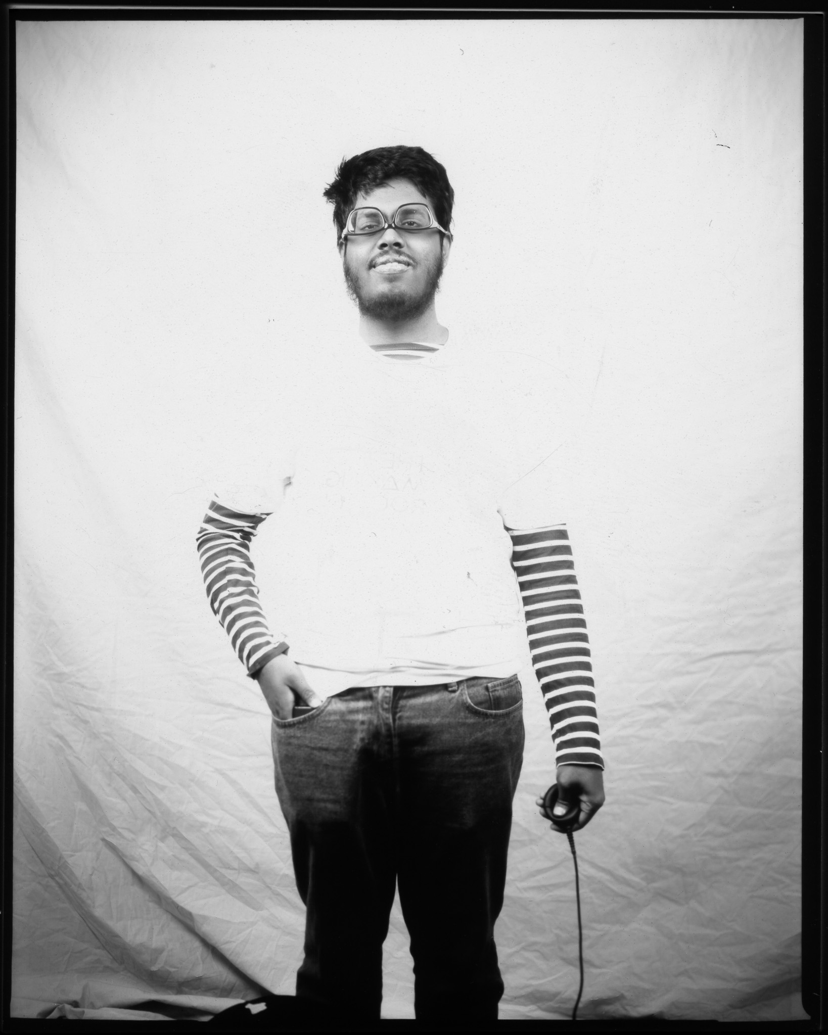 A bearded person stands against a white backdrop with glasses worn upside down, wearing striped sleeves and holding a remote in one hand in a black and white photo.