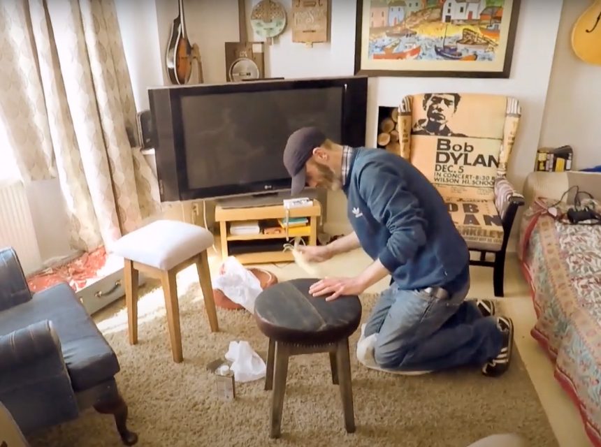 A person kneels on a carpeted floor in a cosy living room while sanding a round wooden stool. The room has a TV, posters, a guitar on the wall, a sofa, and various pieces of furniture.