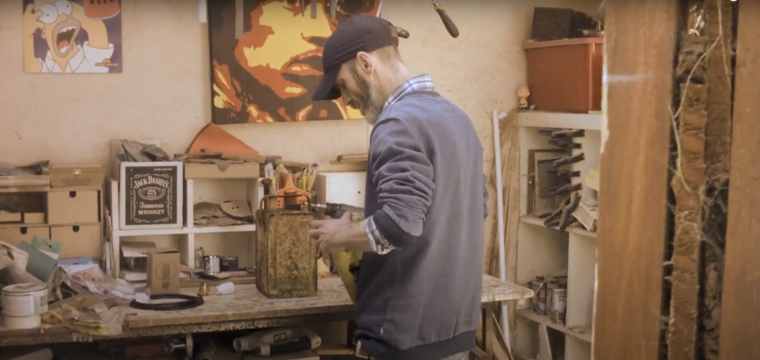 A man wearing a cap and blue jumper works on a wooden object at a cluttered workbench in a workshop, surrounded by tools and various materials, with artwork and shelves visible in the background.