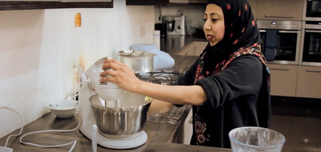 A woman wearing a black headscarf and floral apron uses a stand mixer in a modern kitchen, with pots on the hob and baking ingredients on the worktop.