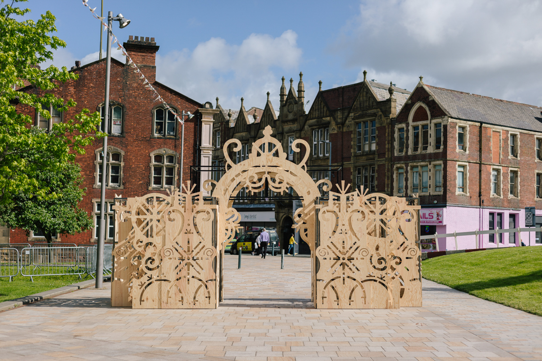 In a public square, a wooden archway by Tim Denton stands. It is made of timber sheets with various ornate and intricate cut outs and shapes.