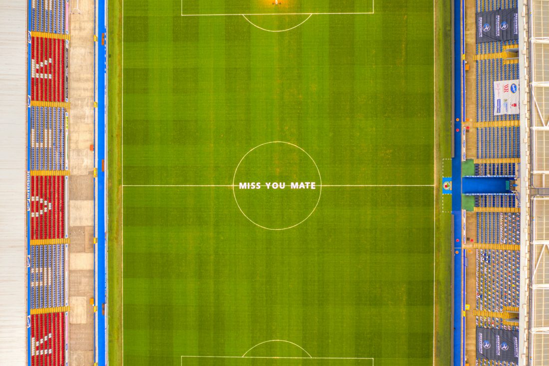 Aerial view of an Blackburn rovers stadium, with “MISS YOU MATE” written in large white letters at the centre of the green pitch.