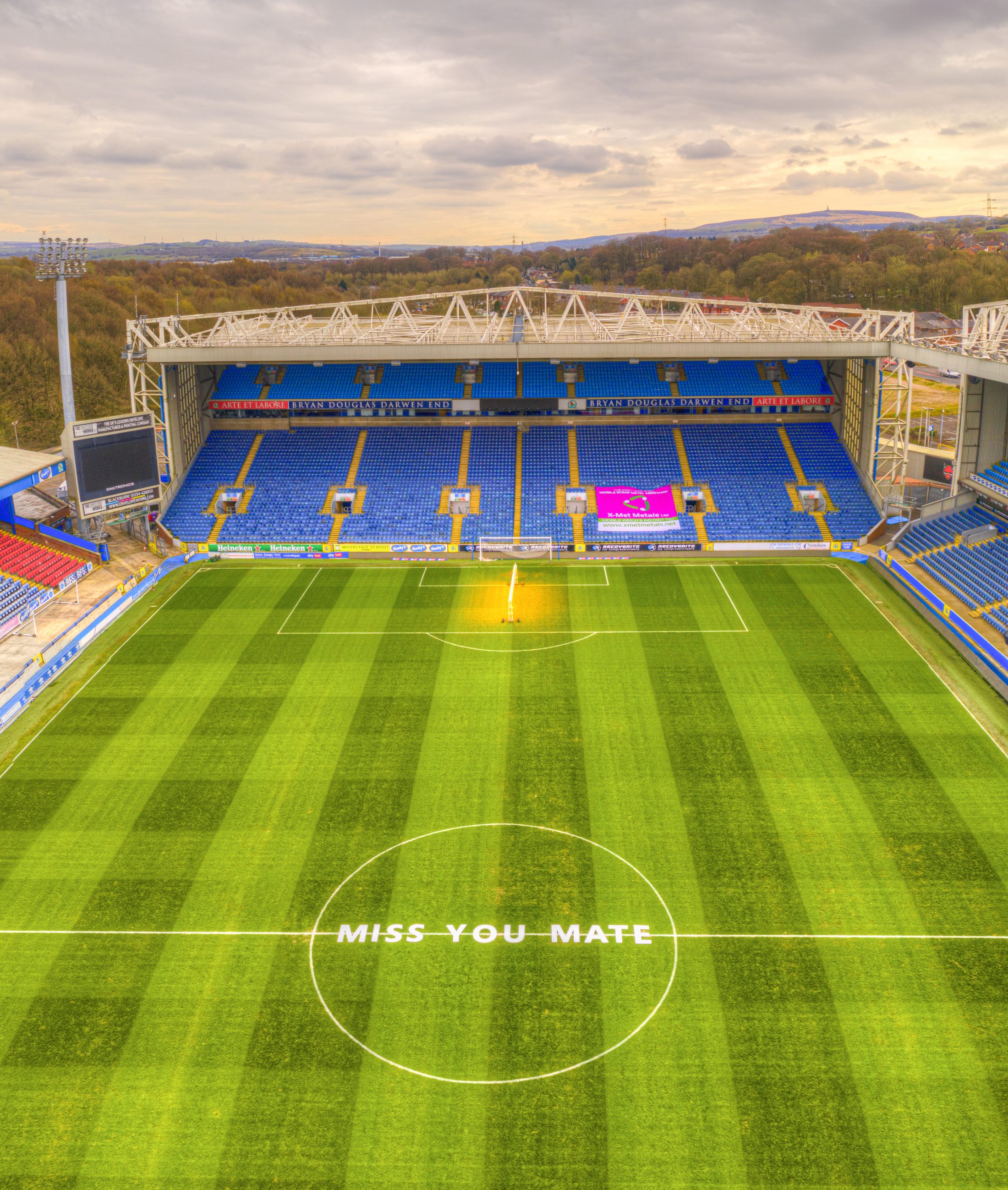 A wide view of an Blackburn Rovers stadium with blue seats and a message on the pitch that reads MISS YOU MATE inside the centre circle.