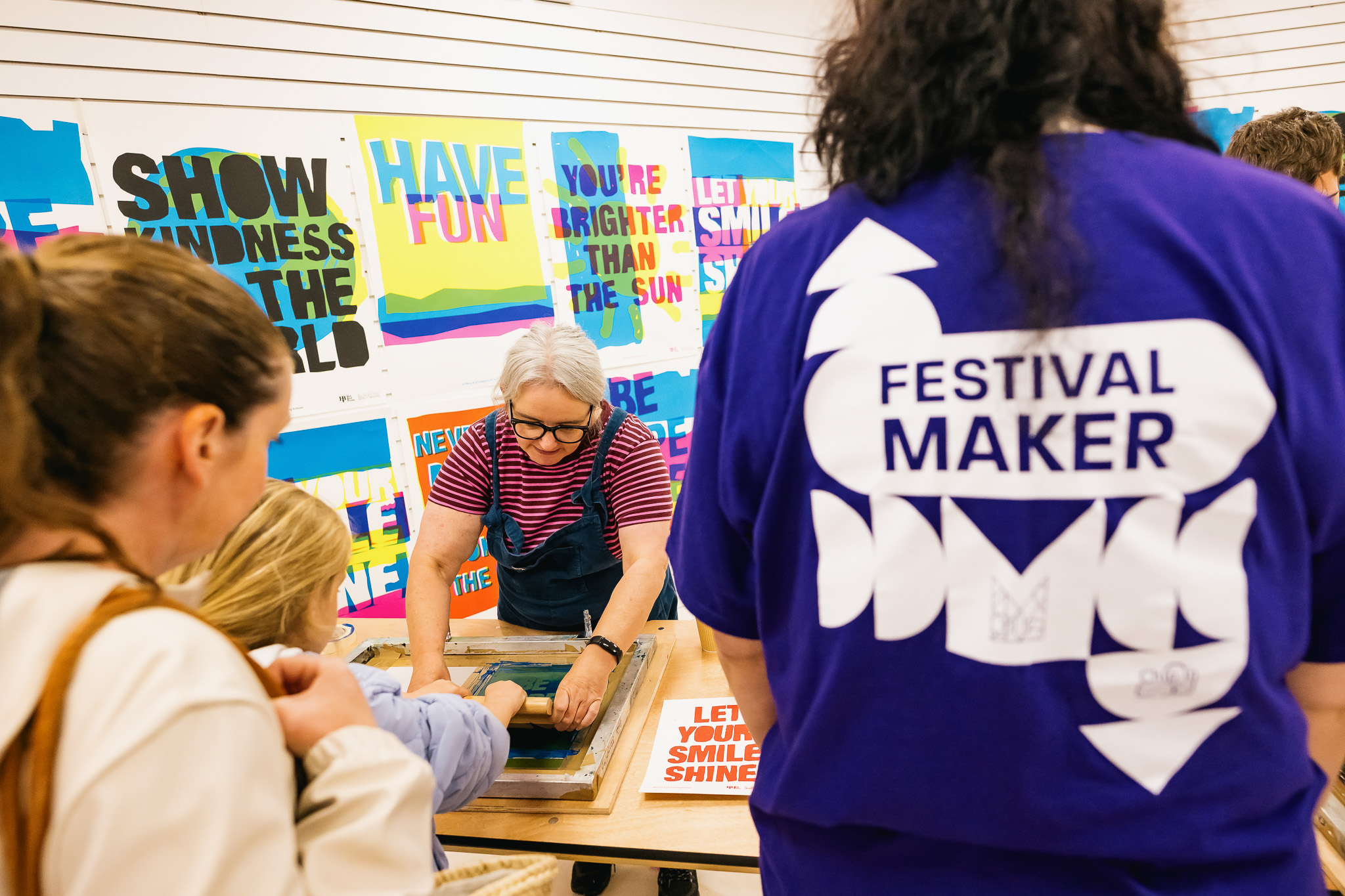 A woman helps a child with a screen printing activity at a table, surrounded by colourful posters with positive messages. In the foreground, a person wears a purple Festival Maker shirt. Other people watch the activity.