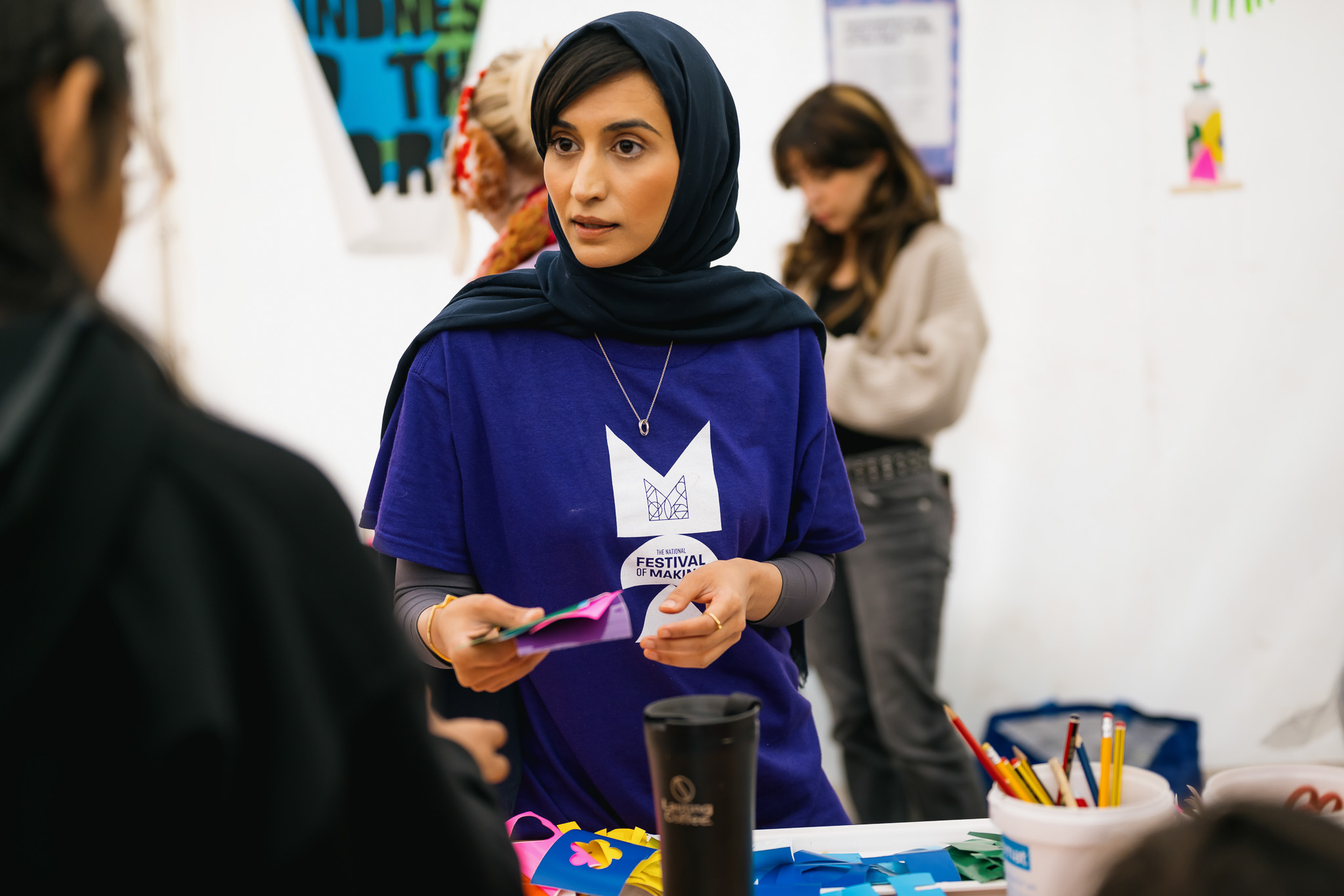 A woman in a navy hijab and purple Festival Maker shirt talks to someone at a table with colourful arts and crafts supplies. People and posters are visible in the background.