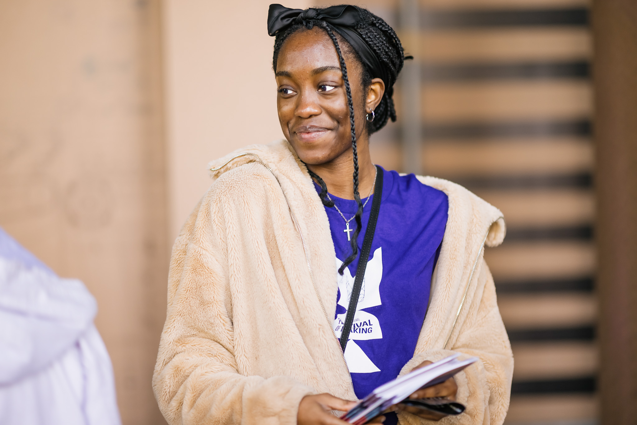 A young woman with plaited hair, wearing a black headband, a beige faux fur coat, and a purple T-shirt, smiles while holding a notebook and pen. She stands indoors with a blurred background.