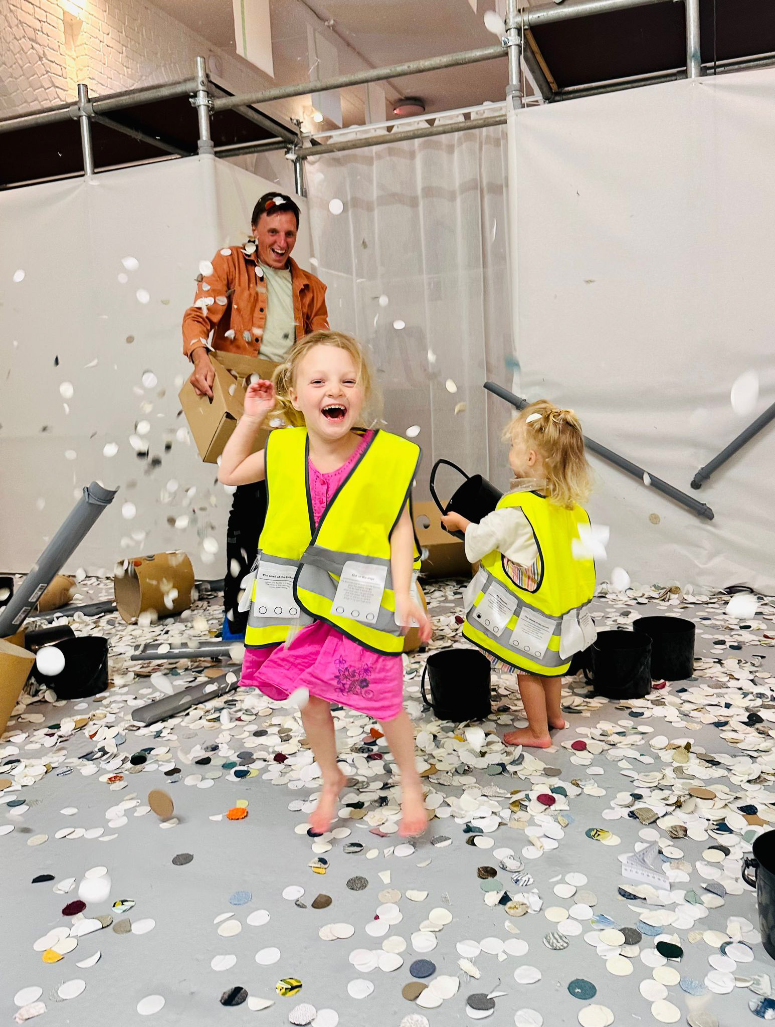 Two young children in reflective vests play joyfully among scattered paper discs, with an adult smiling in the background. The floor is covered in confetti-like paper, creating a playful, messy scene indoors.