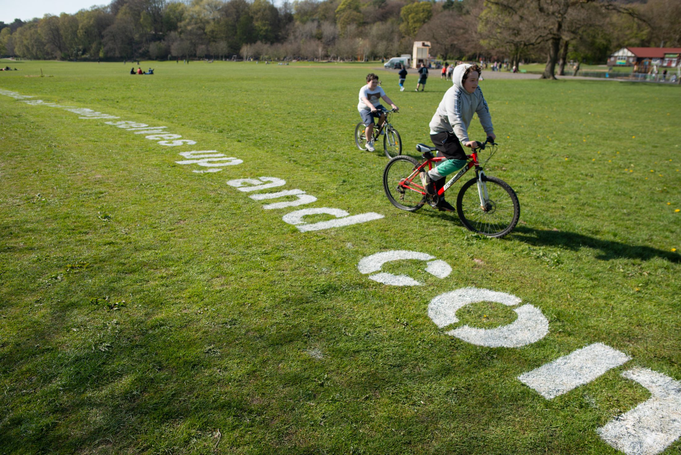 Two people cycle on a grassy field with large painted letters on the grass. Trees and a building are visible in the background, suggesting a park setting.