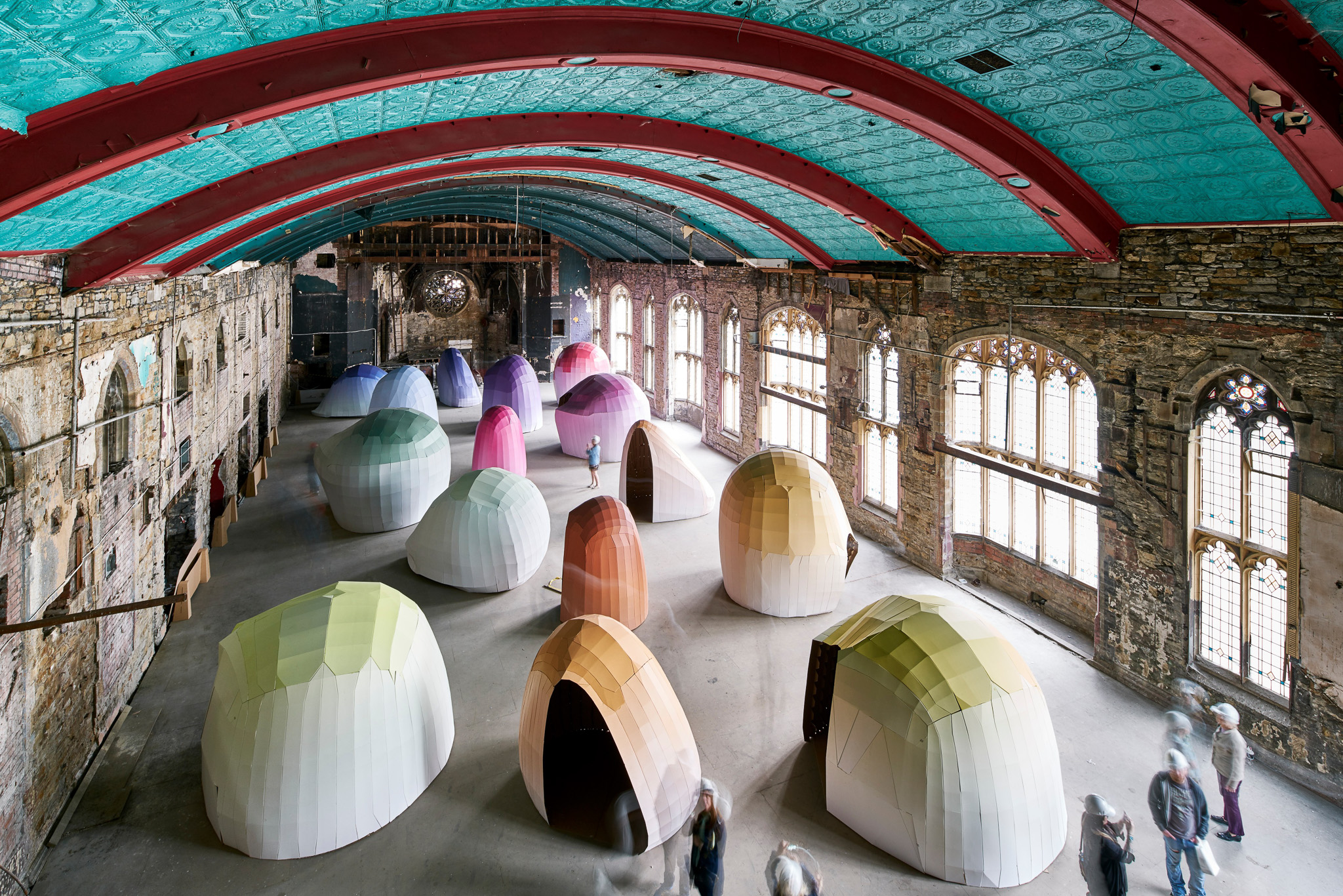 Large, colourful, cardboard dome-shaped structures are arranged inside a spacious, old building with tall arched windows, exposed stone walls, and a turquoise ceiling with red beams. Several blurred figures walk among the domes.