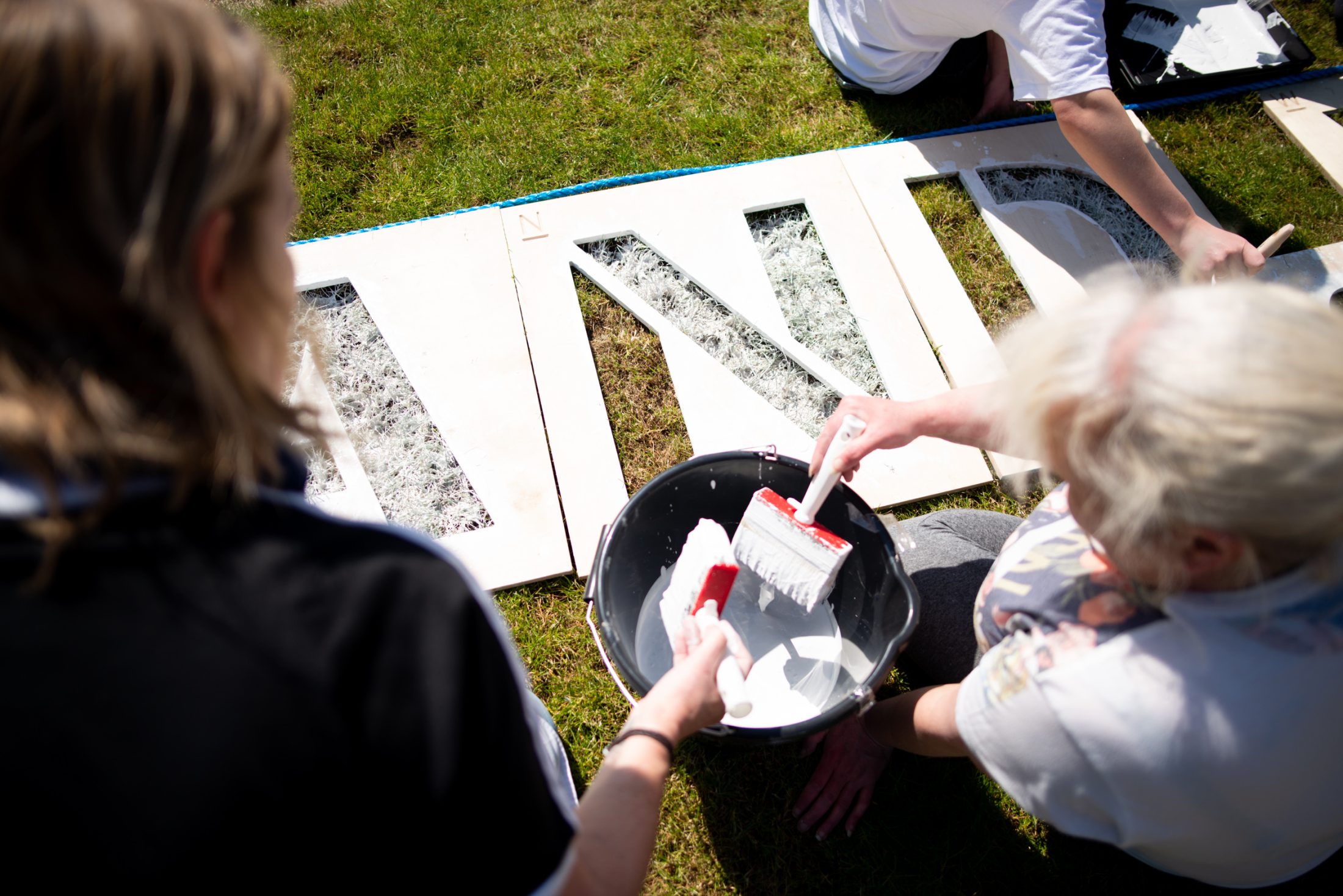 Two people, seen from above, dip paint rollers into a bucket of white paint while painting large stencilled letters on grass in sunny outdoor conditions.