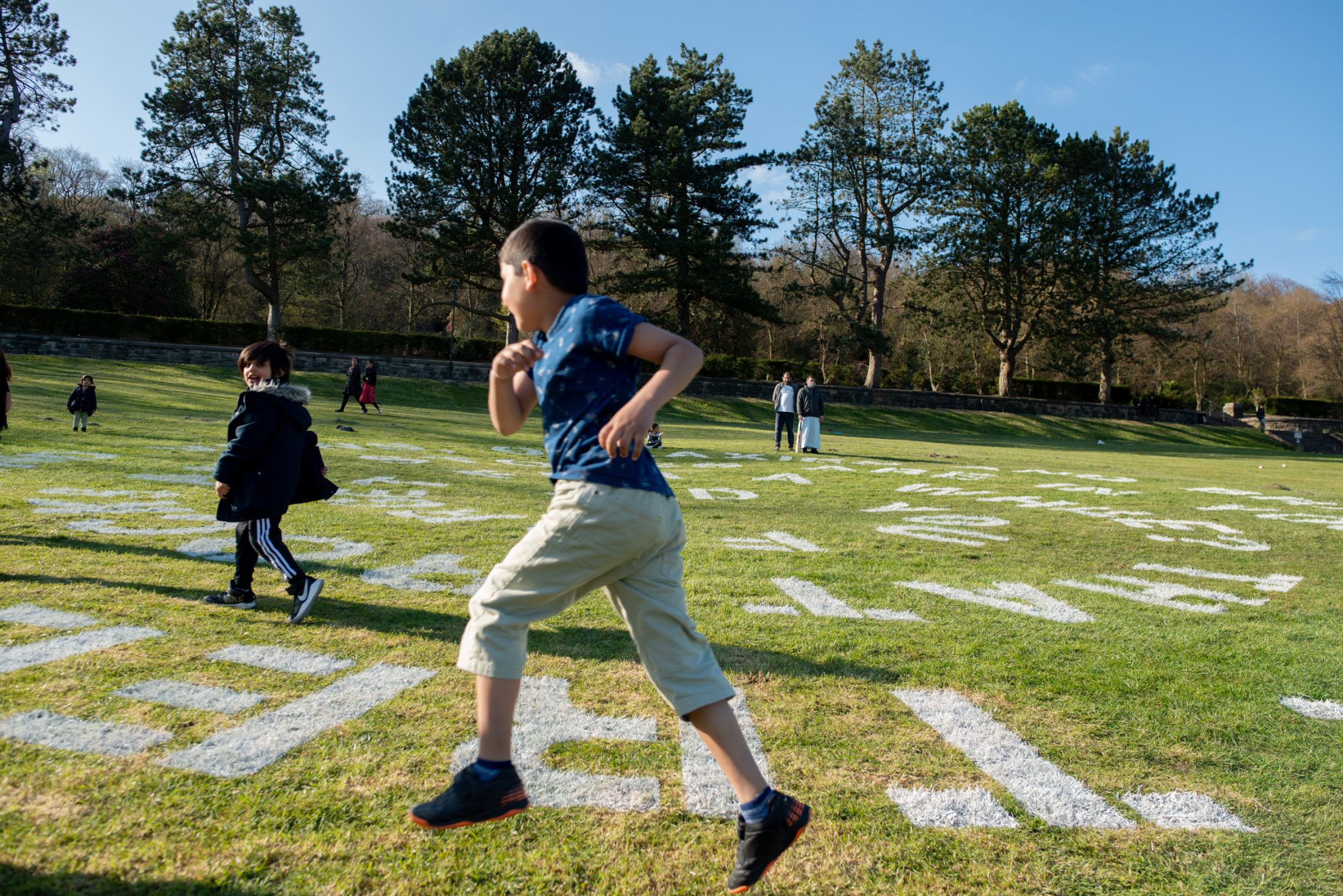 Two boys run and play on a grassy field with large white letters painted on it. More people are visible in the background, with tall trees and a clear blue sky overhead.