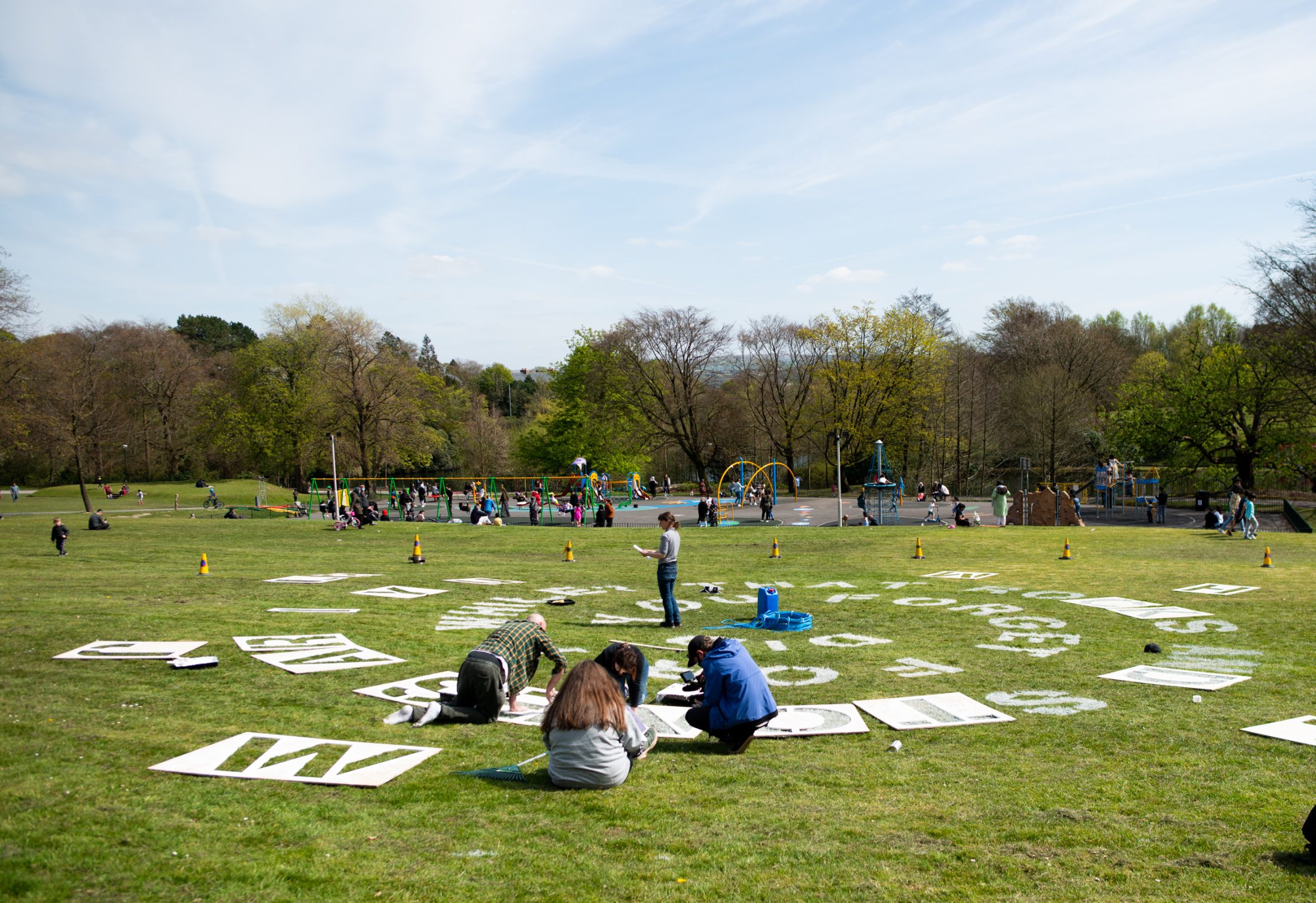 People work on large letter boards spread out on grass in a park, with children playing on playground equipment in the background. Trees and blue sky frame the lively outdoor scene.
