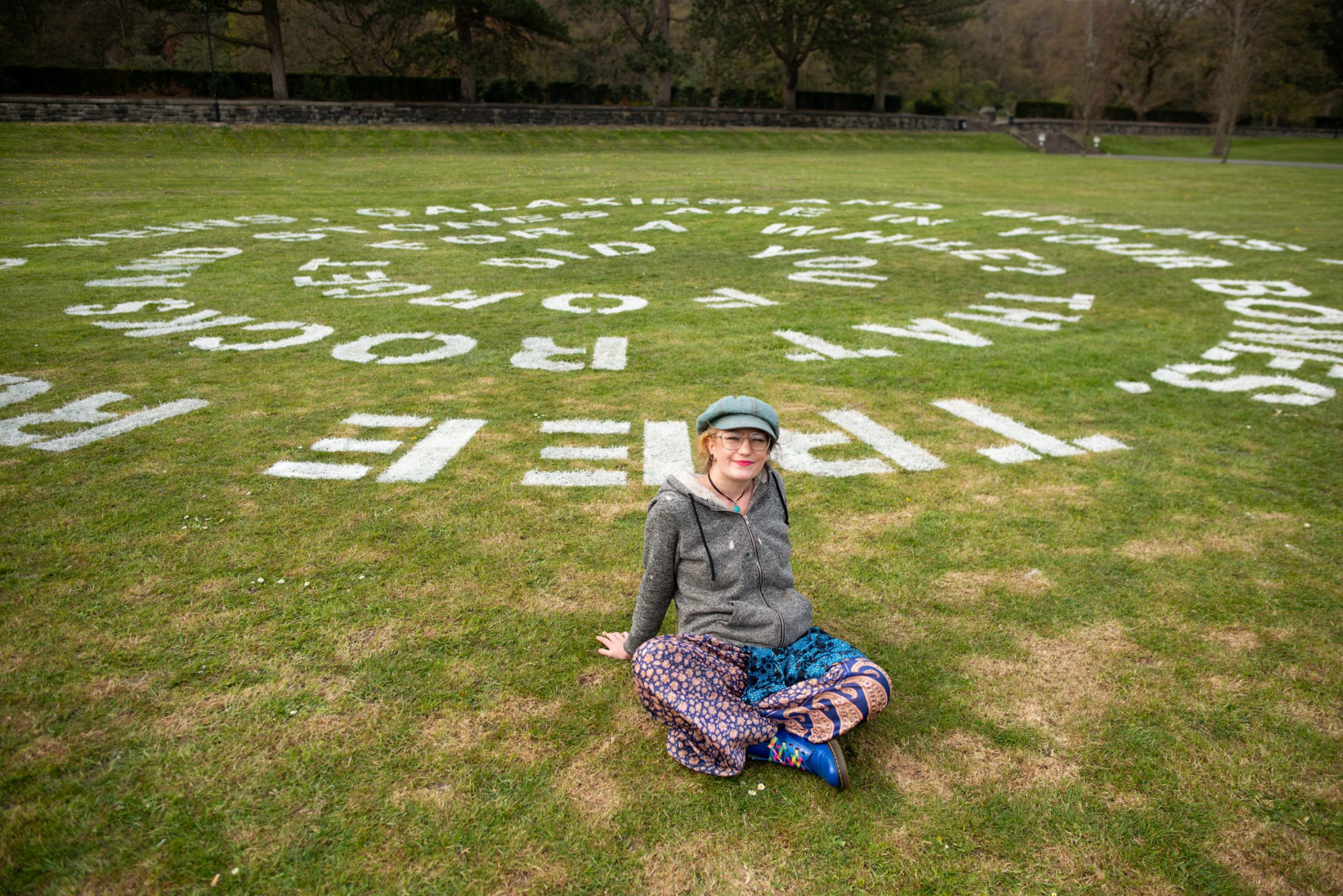 A person with glasses and patterned trousers sits on grass, smiling, in front of large white words painted in a spiral pattern on the lawn in a park-like setting. Trees and a stone wall are in the background.