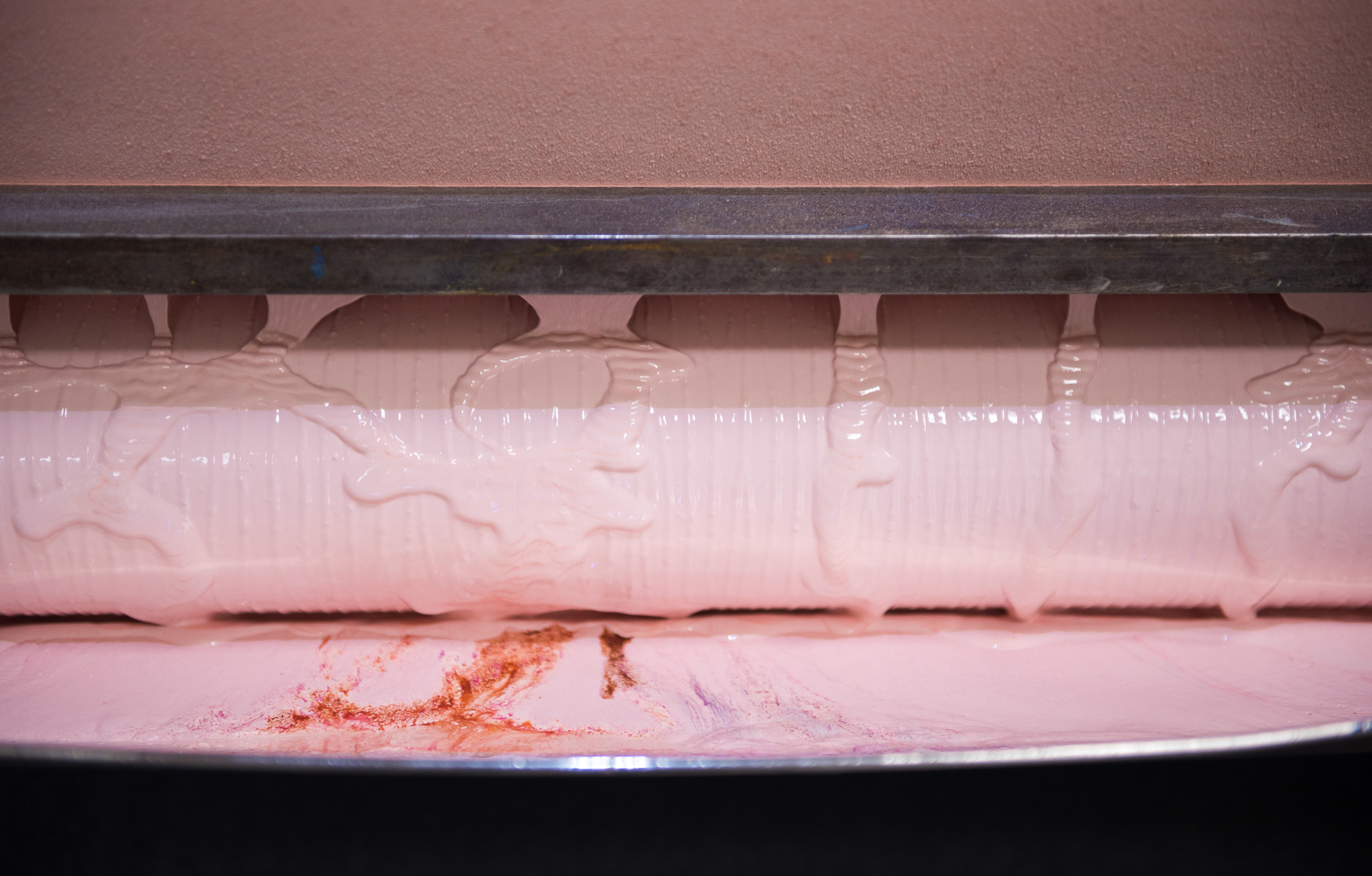 A close-up of a large industrial roller coated with thick, pale pink liquid and some reddish streaks, likely in a manufacturing or food processing setting.
